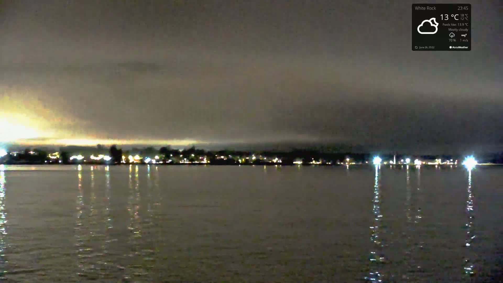 At night under a mostly cloudy sky, the lights of a town are reflected on a calm body of water.