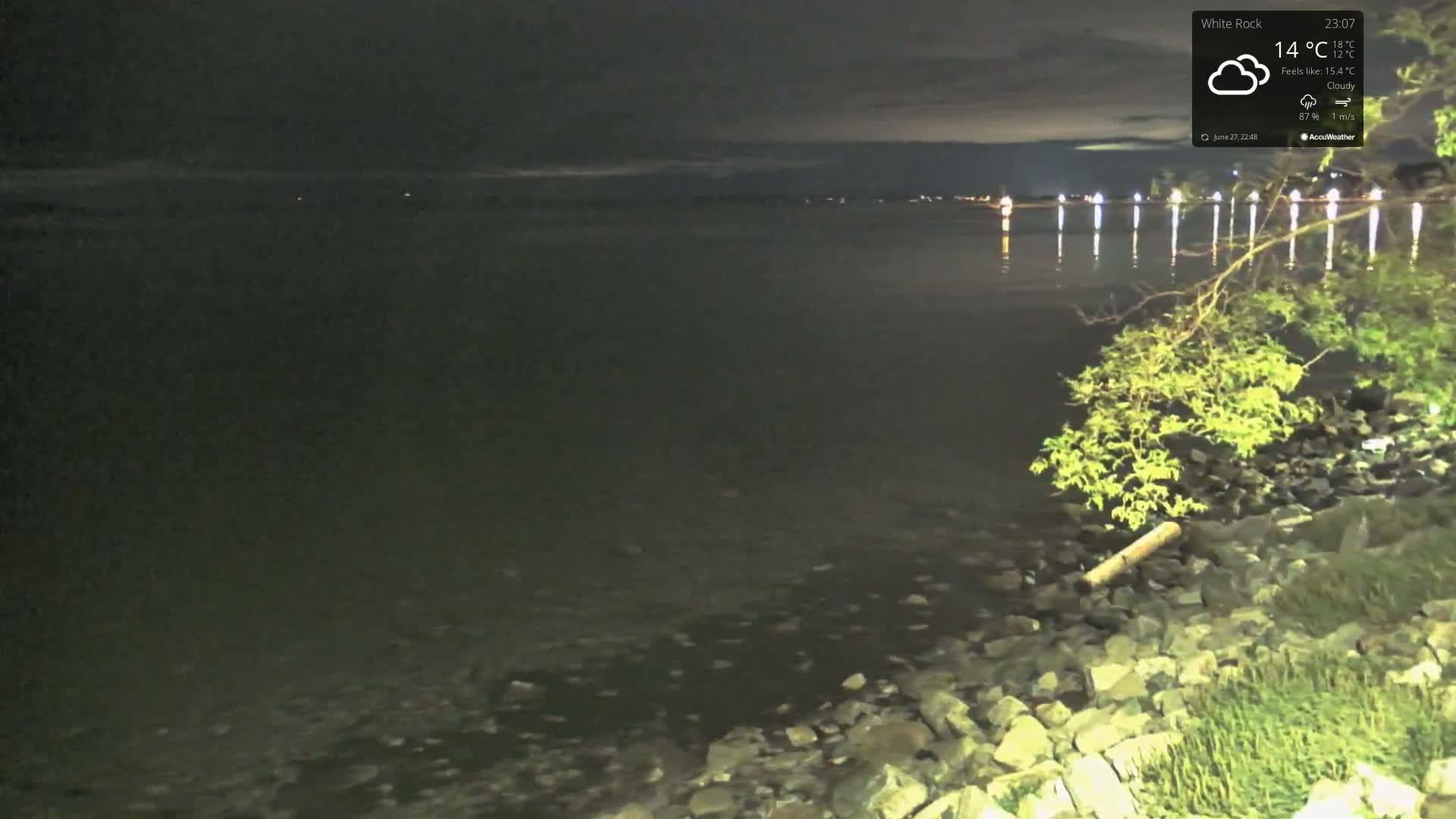 A nighttime view of a dark, calm body of water with lights visible in the distance under a cloudy sky.