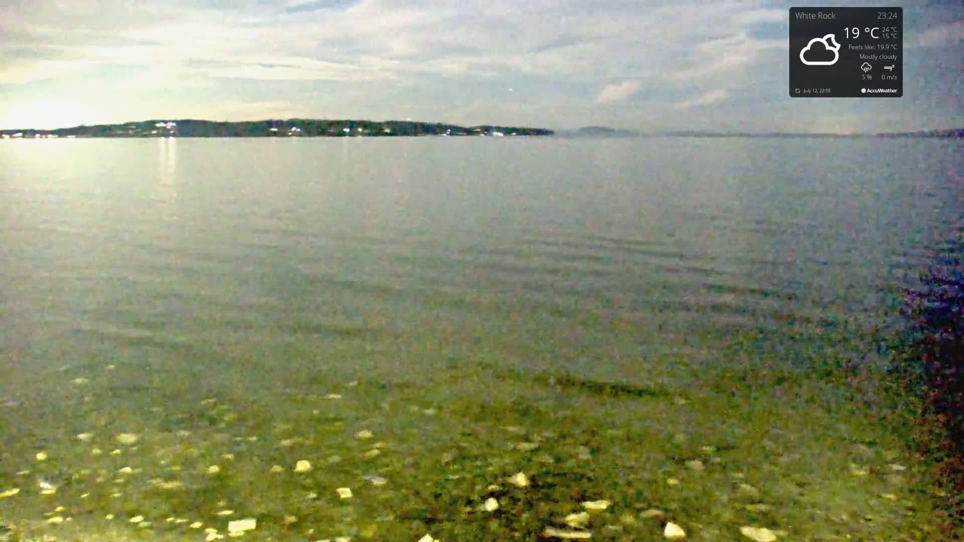 A mostly cloudy night scene shows a calm body of water with a distant shoreline under a pale sky.