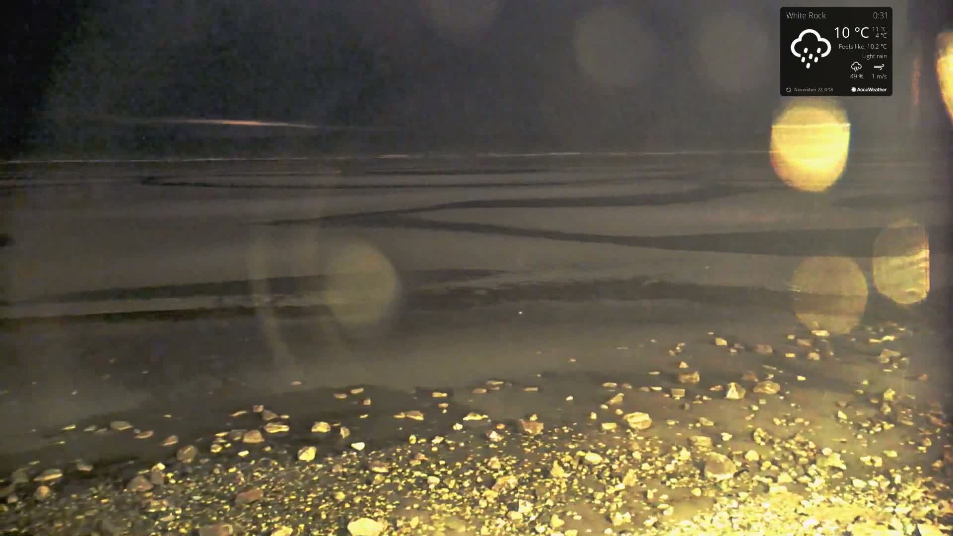 A dark, rainy night view reveals a wet, rocky shoreline illuminated by warm artificial lights creating prominent lens flares, with a reflective expanse of water or wet sand displaying faint patterns stretching towards the horizon.