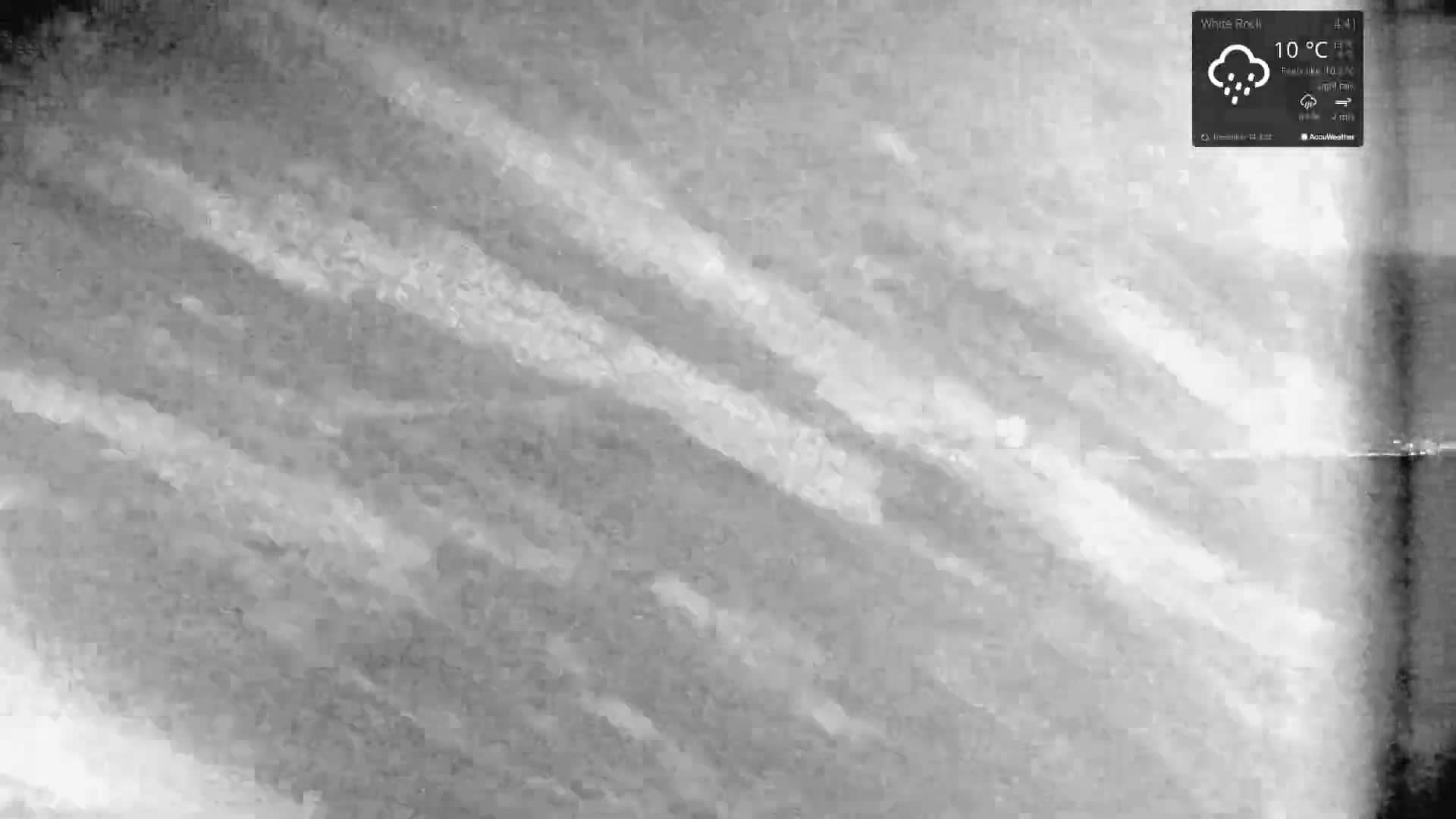 A large, bare tree adorned with bright white string lights stands prominently next to a paved pathway and railway tracks, overlooking a calm, dark body of water with faint distant lights under a mostly cloudy night sky.