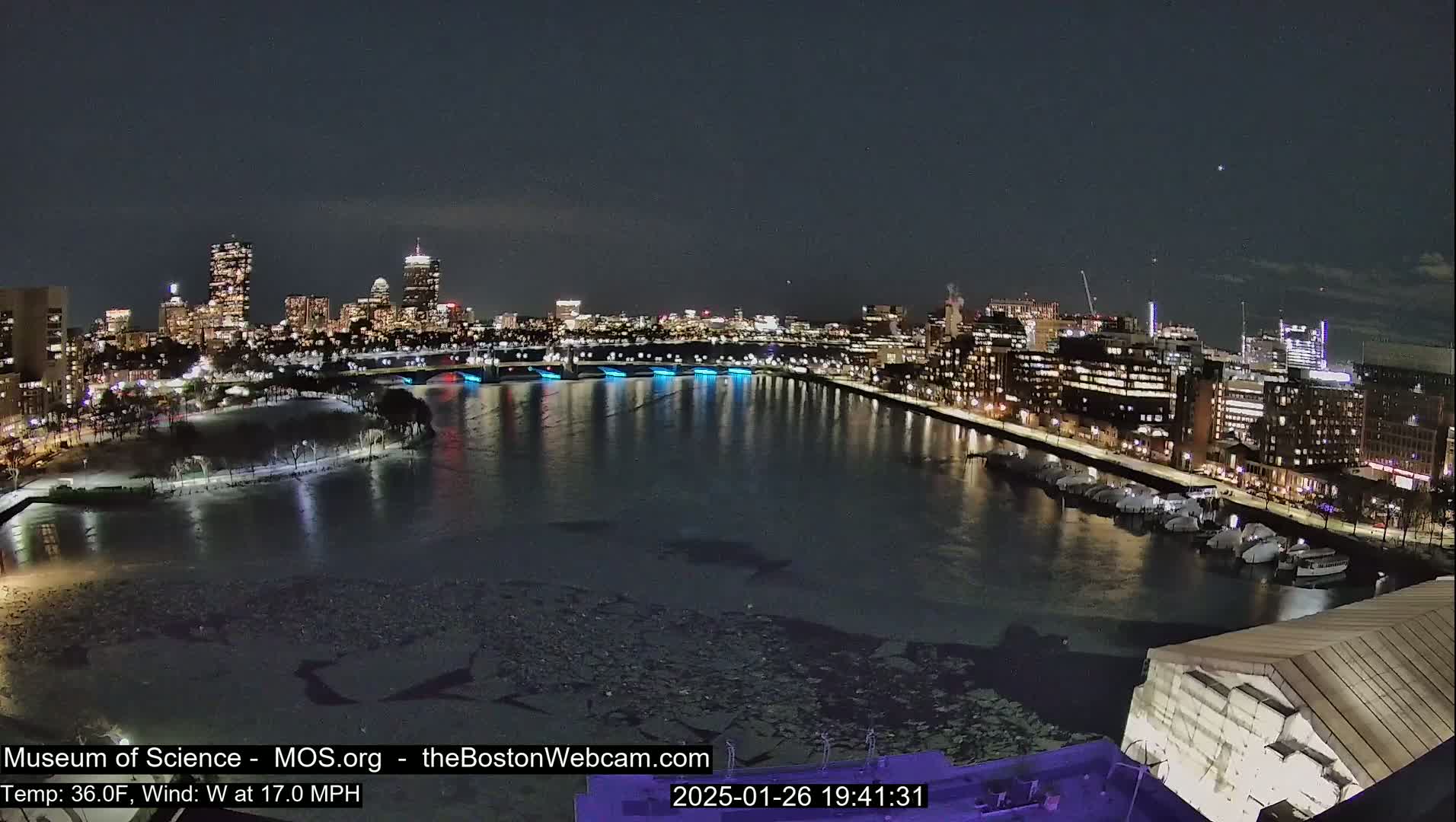 A nighttime view of a city skyline across a partially ice-covered river, with a 36°F temperature and a 17 mph west wind.