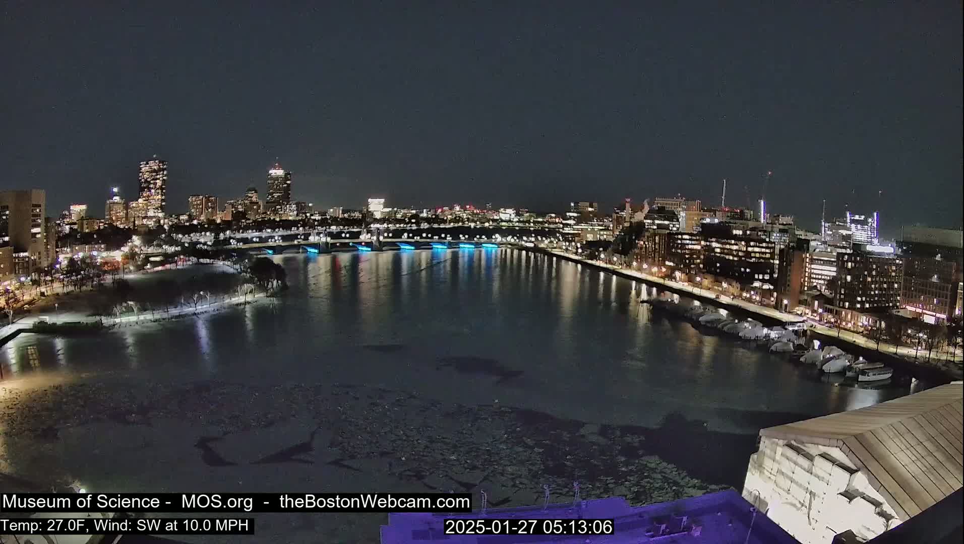 A nighttime view of a city skyline across a partially ice-covered river, with a southwest wind at 10 mph and a temperature of 27 degrees Fahrenheit.