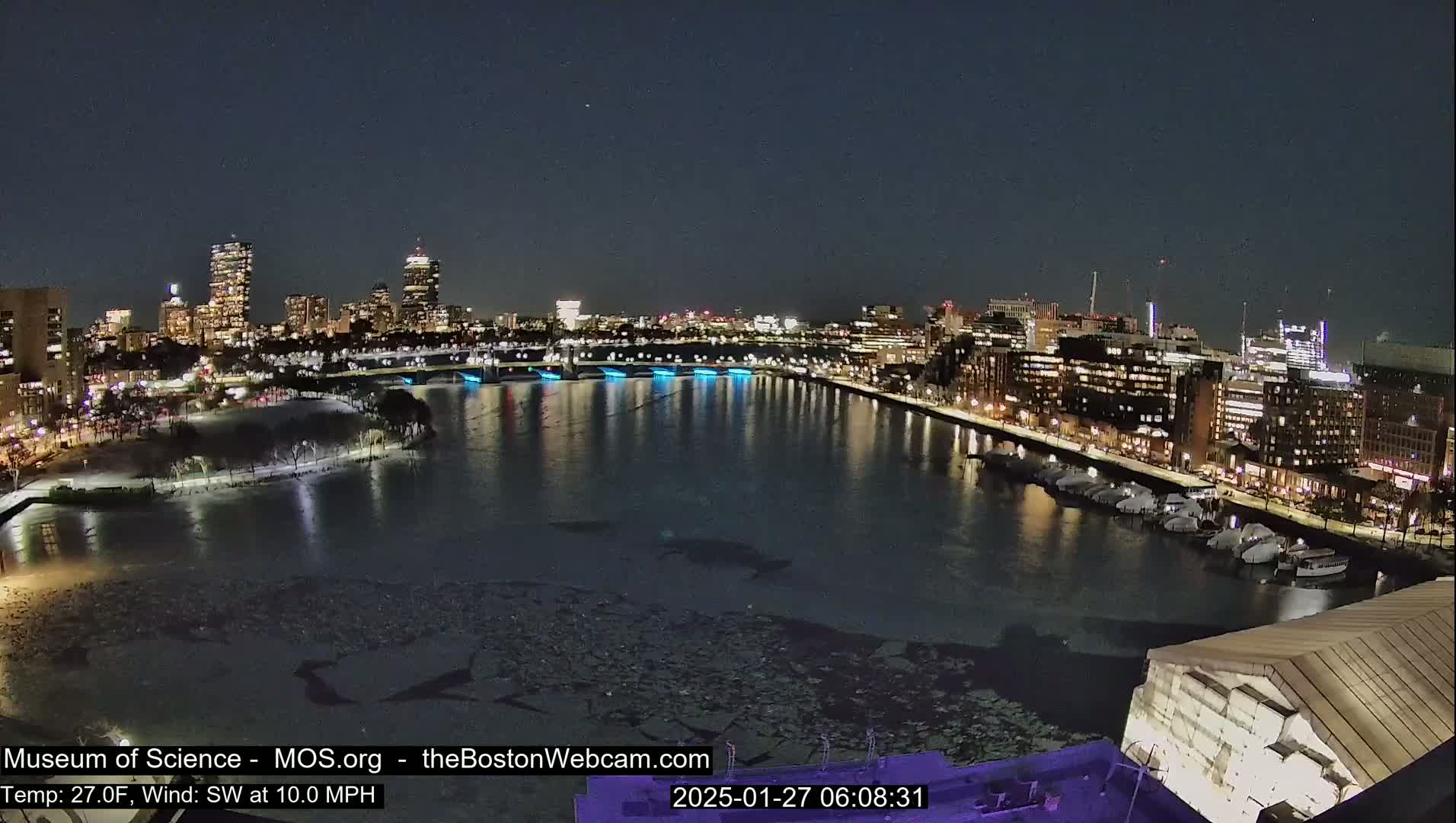 A nighttime view of a city skyline across a partially ice-covered river, with a southwest wind at 10 mph and a temperature of 27°F.