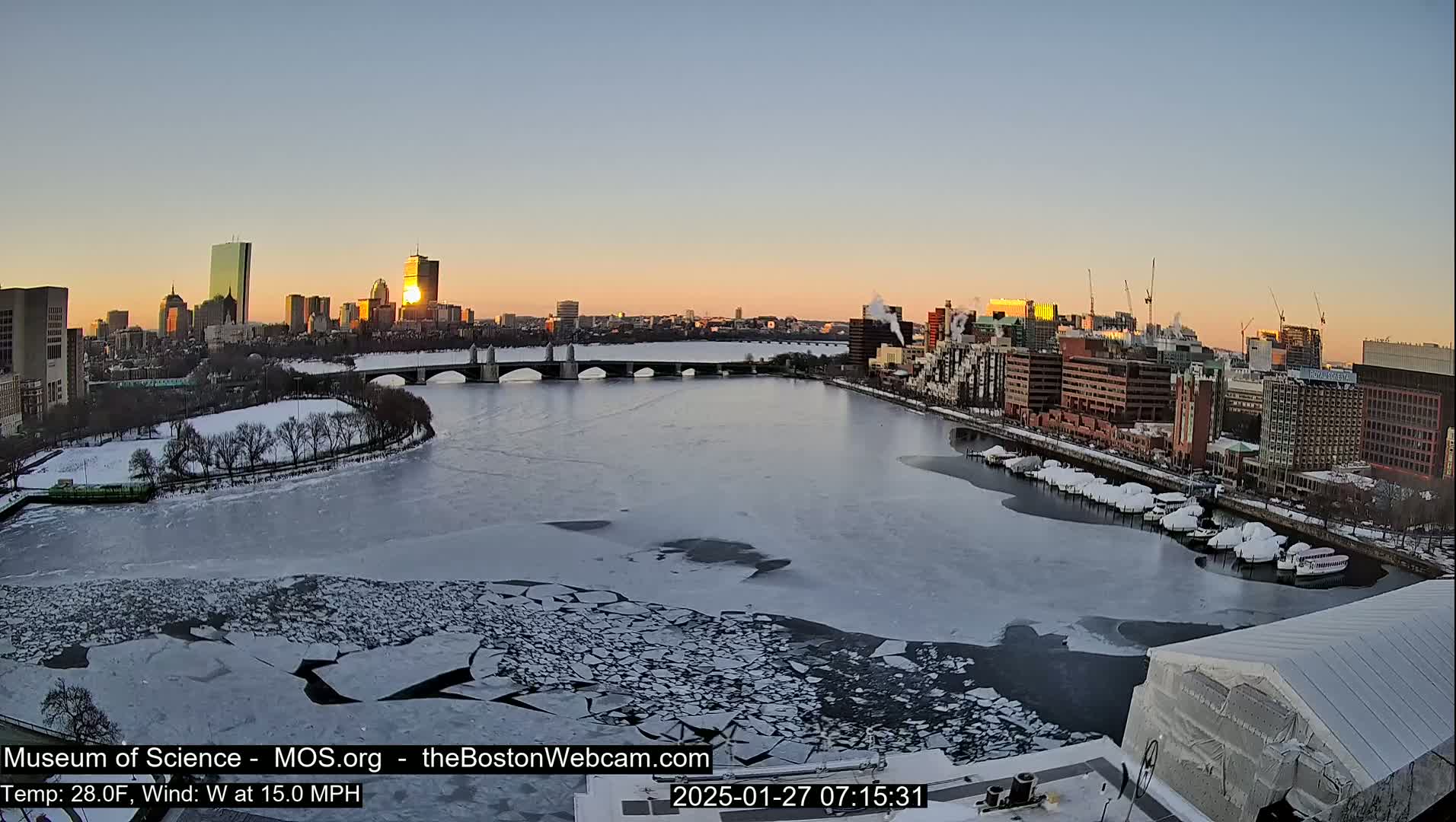 A partially frozen river flows through a city at sunrise, under a clear sky with a cold wind.