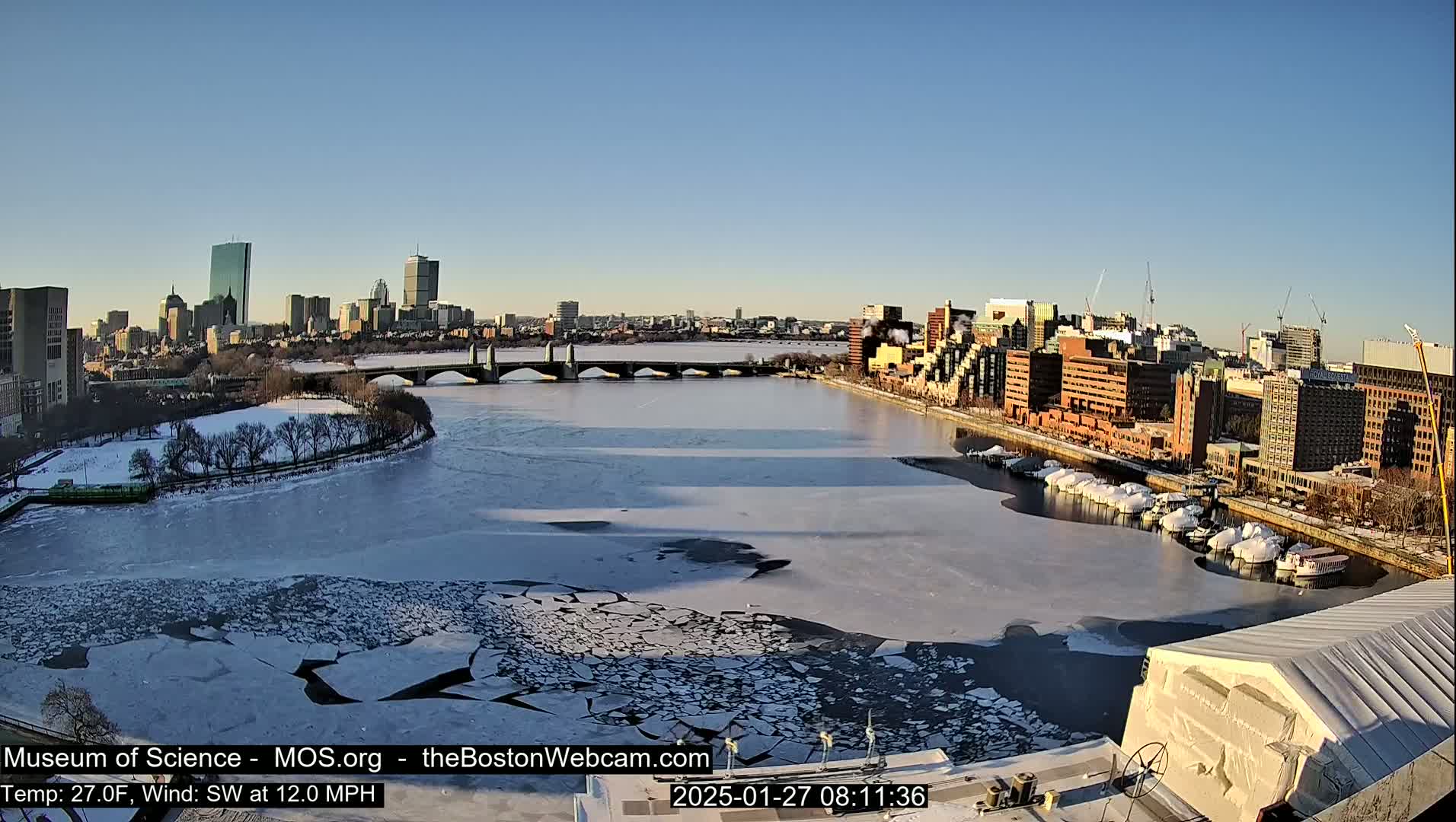A partly frozen river flows through a city, with a bridge spanning the water under a clear blue sky.