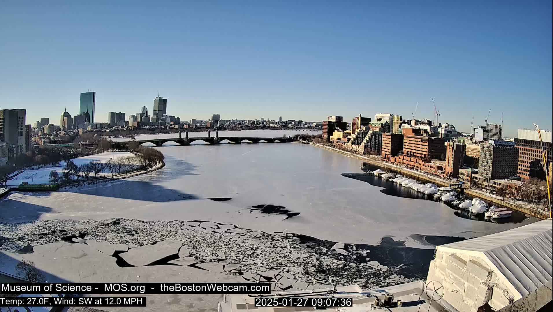 A partly frozen river flows past a city skyline under a clear, sunny sky.