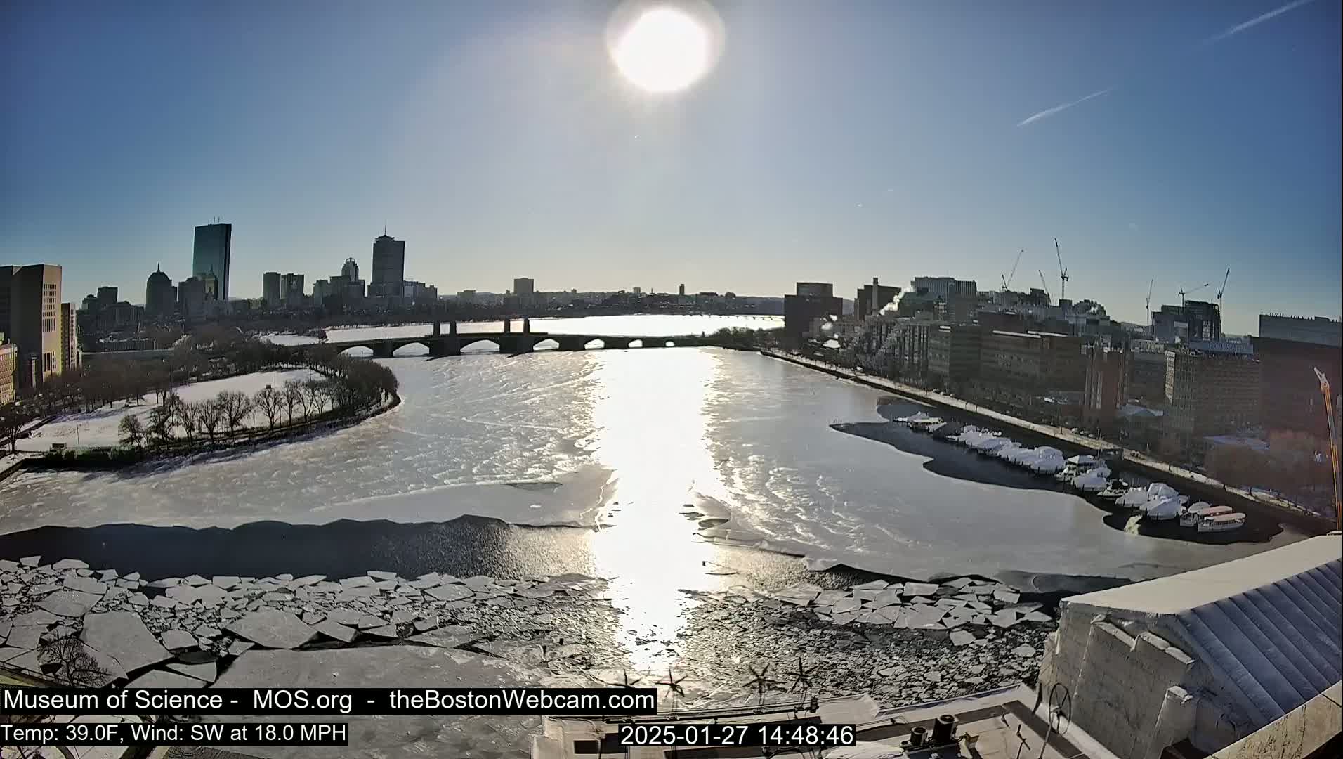 A sunny day shows a partly frozen river flowing through a city, with broken ice floes scattered on the water's surface and a bridge spanning the waterway.