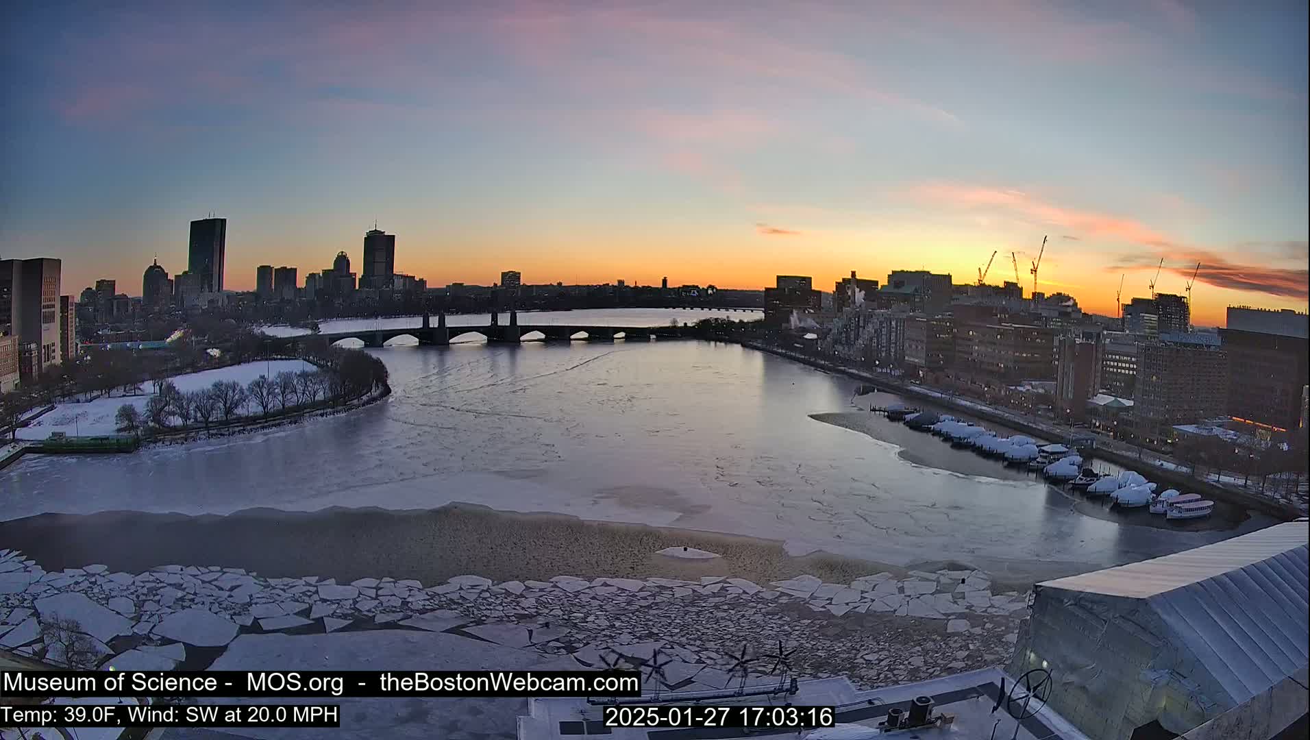 A partially frozen river flows past a city skyline at sunset, with ice floes scattered across the water's surface.
