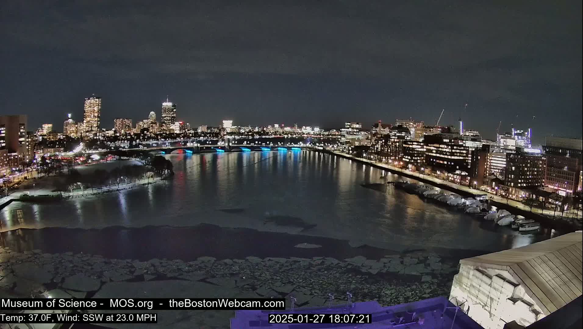 A nighttime cityscape with a river partially covered in ice, under a dark sky with a strong south-southwesterly wind.