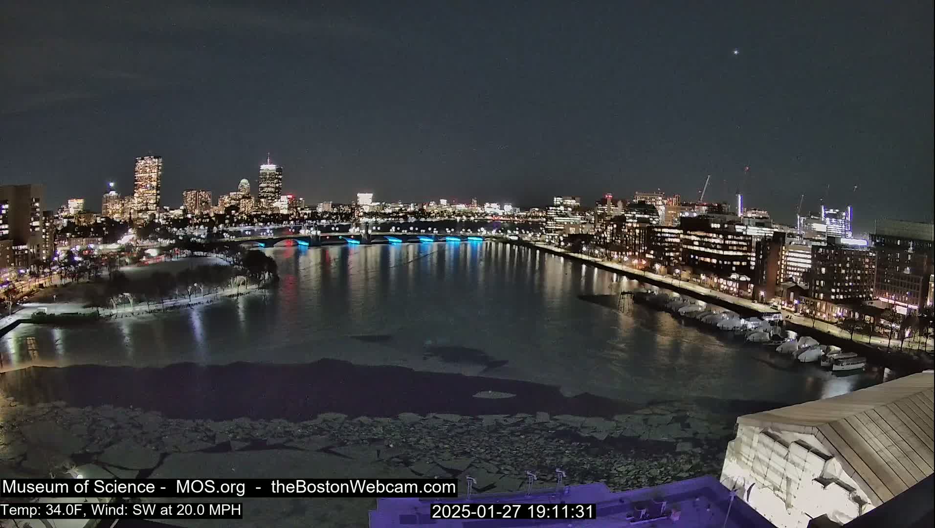 A nighttime view of a city skyline across a partially frozen river, under a clear sky with a southwest wind at 20 mph and a temperature of 34°F.