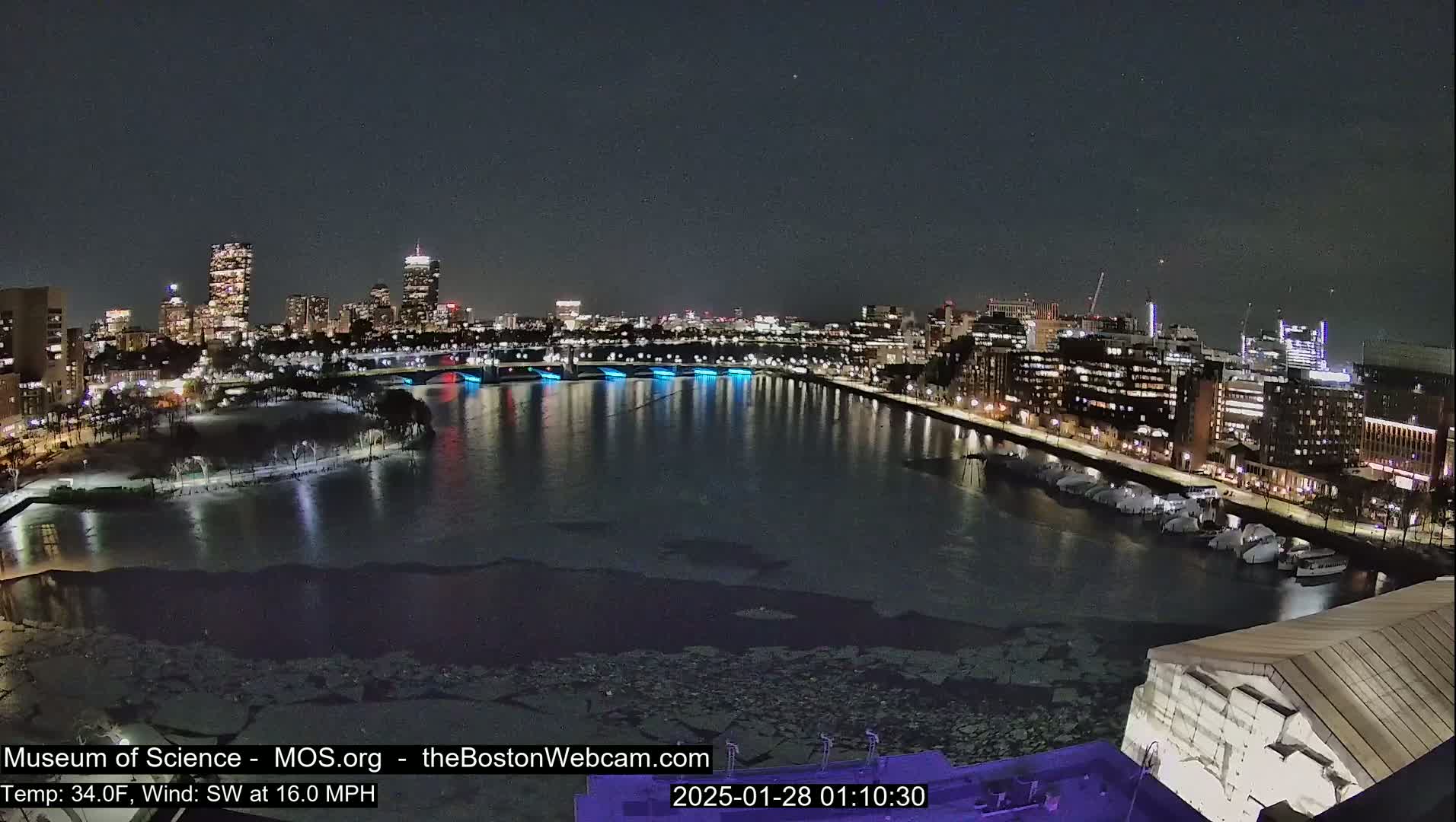 A nighttime cityscape with a partially frozen river, illuminated buildings, and a southwest wind of 16 mph at a temperature of 34°F.