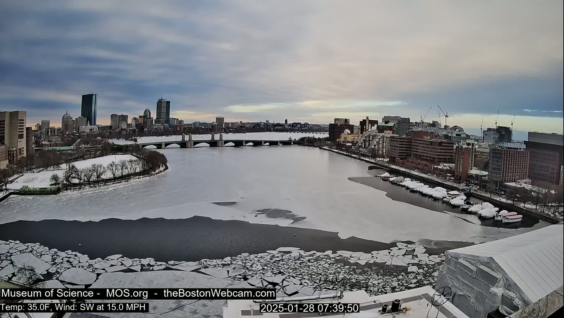 A partially frozen river flows past a city skyline under a cloudy sky, with boats covered in snow docked along the riverbank.