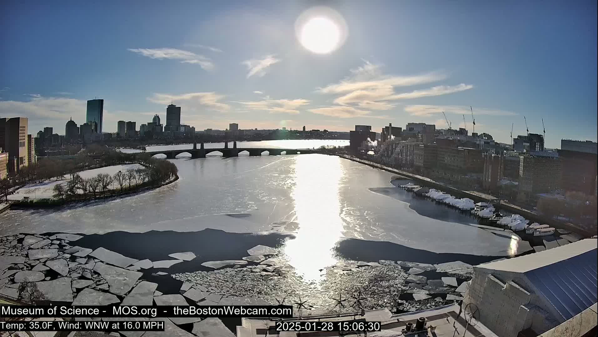 A sunny day shows a partially frozen river flowing past a city skyline with a bridge, boats docked along the riverbank, and snow-covered areas.