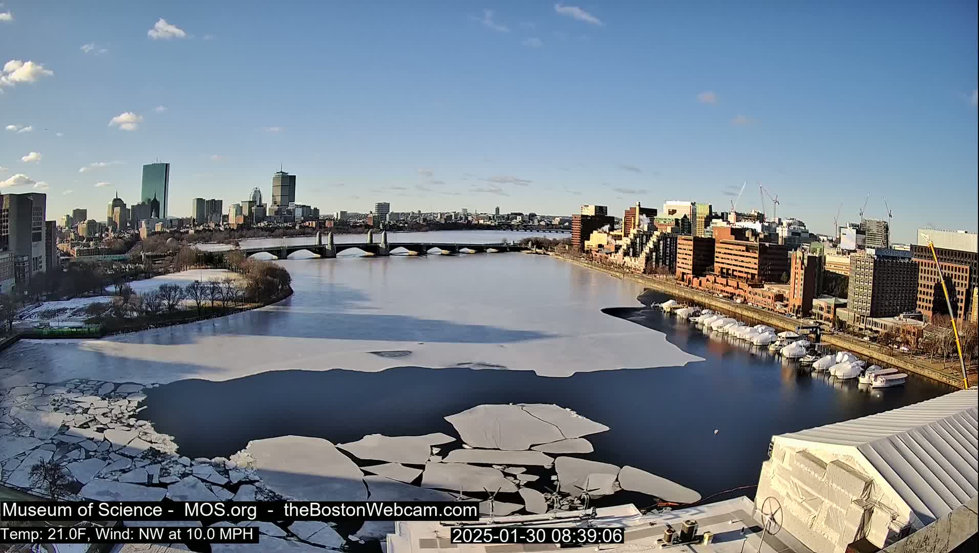 A partially frozen river flows through a city, with a bridge spanning the waterway under a clear, sunny sky.