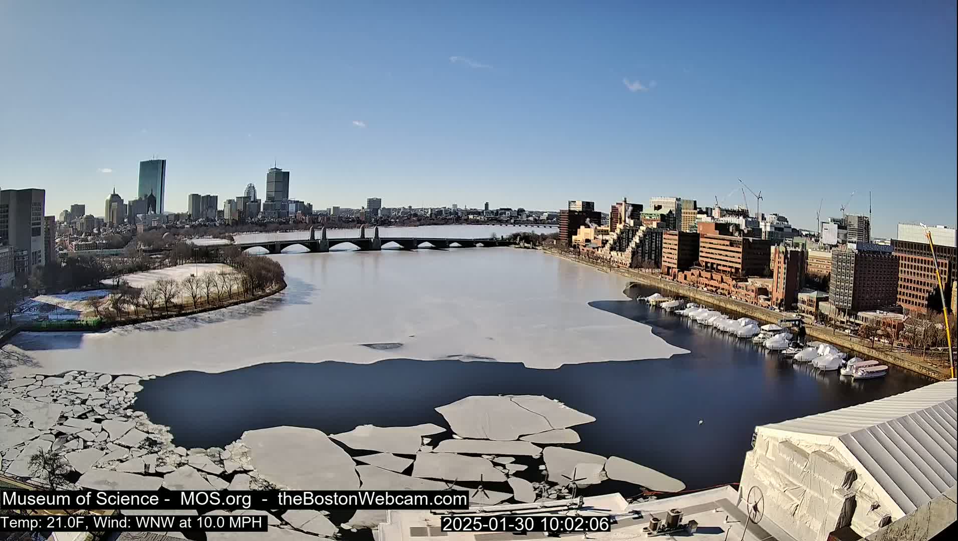 A partially frozen river flows past a city skyline under a clear blue sky, with boats moored along the riverbank.