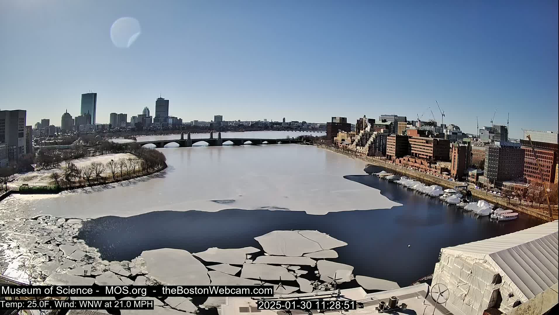 A partly frozen river flows past a city skyline under a clear blue sky.