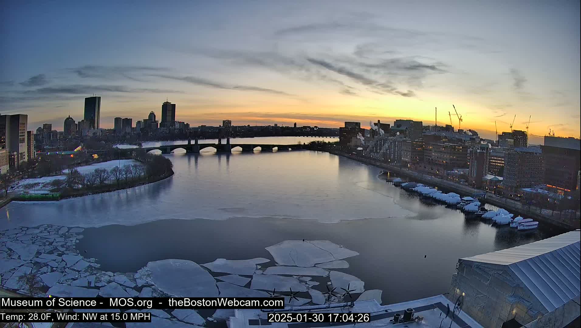 A partly frozen river flows through a city at sunset, with ice floes scattered across its surface under a partly cloudy sky.