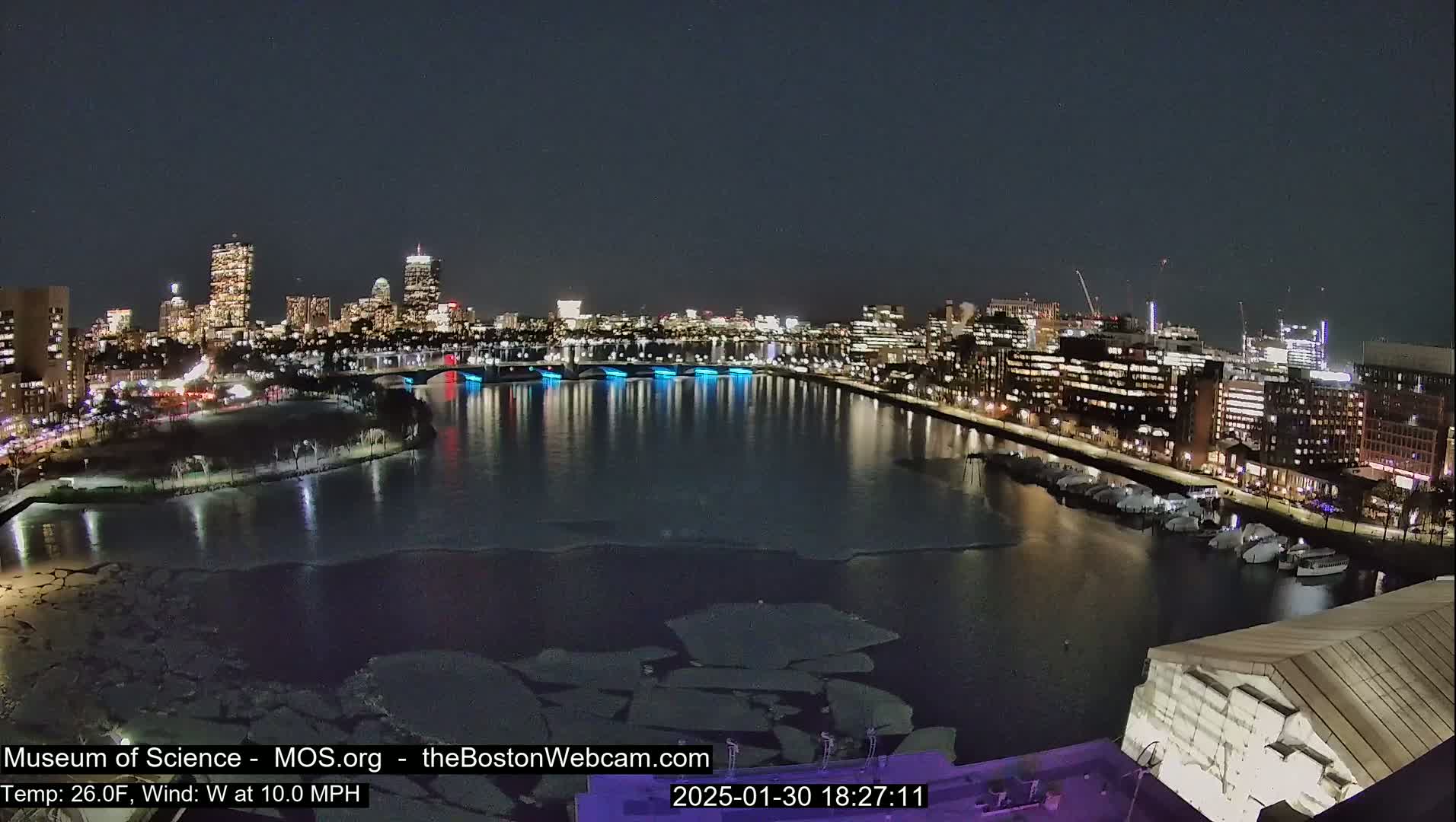 A nighttime view of a city skyline across a partially frozen river, with a 26°F temperature and a 10 mph west wind.