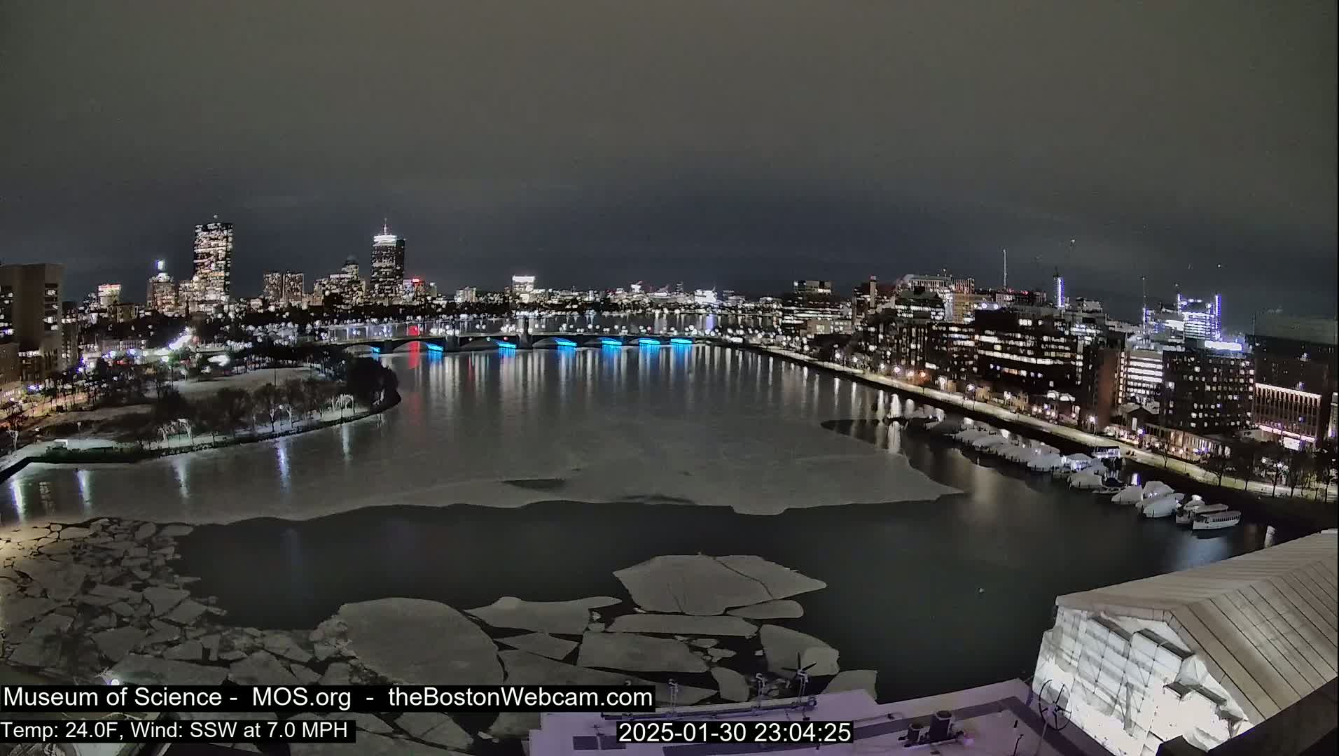 A nighttime cityscape with a partially frozen river, illuminated bridges, and a city skyline under a dark sky.