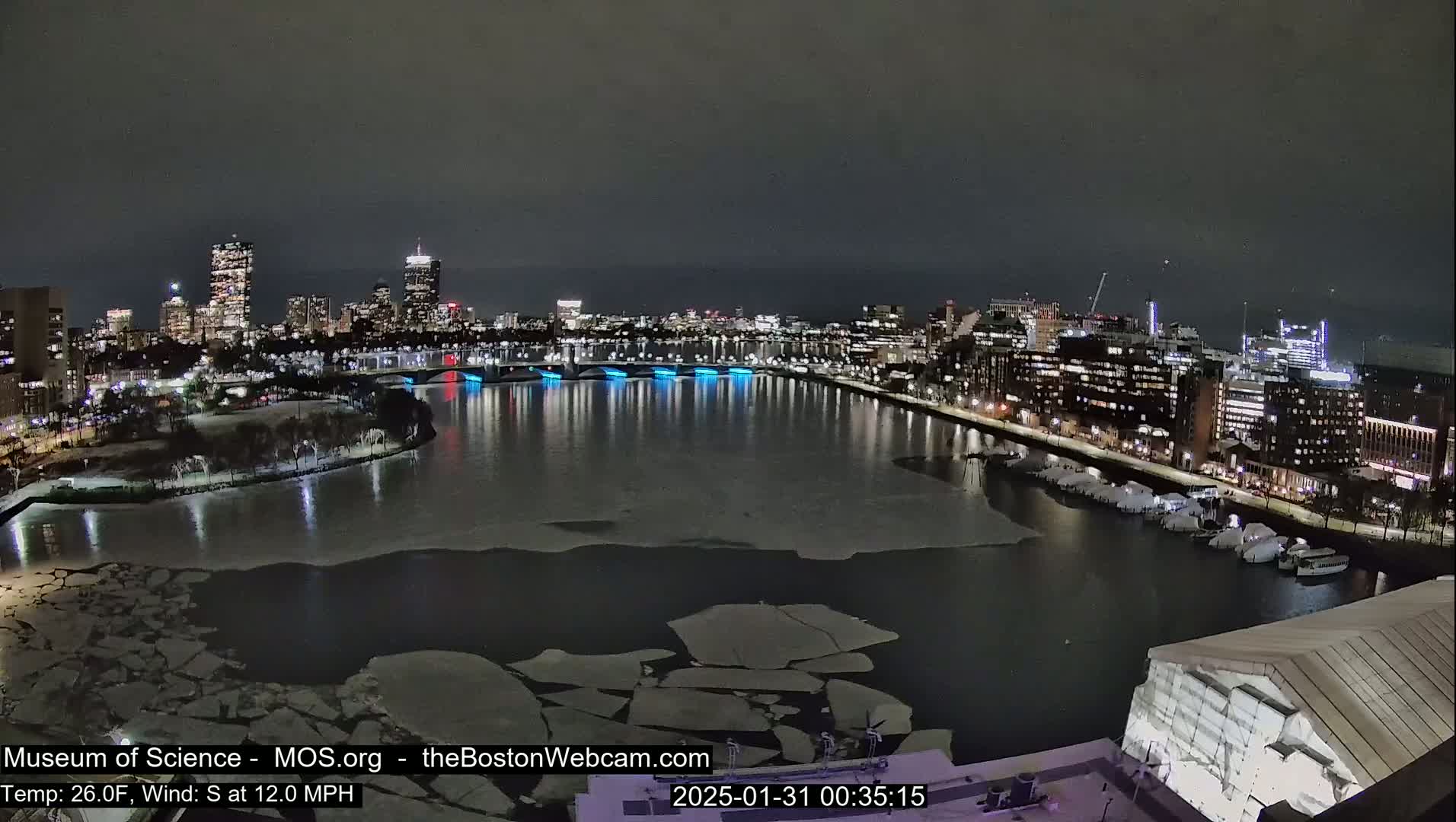 A nighttime view of a city skyline across a partially ice-covered river, with a south wind at 12 mph and a temperature of 26 degrees Fahrenheit.
