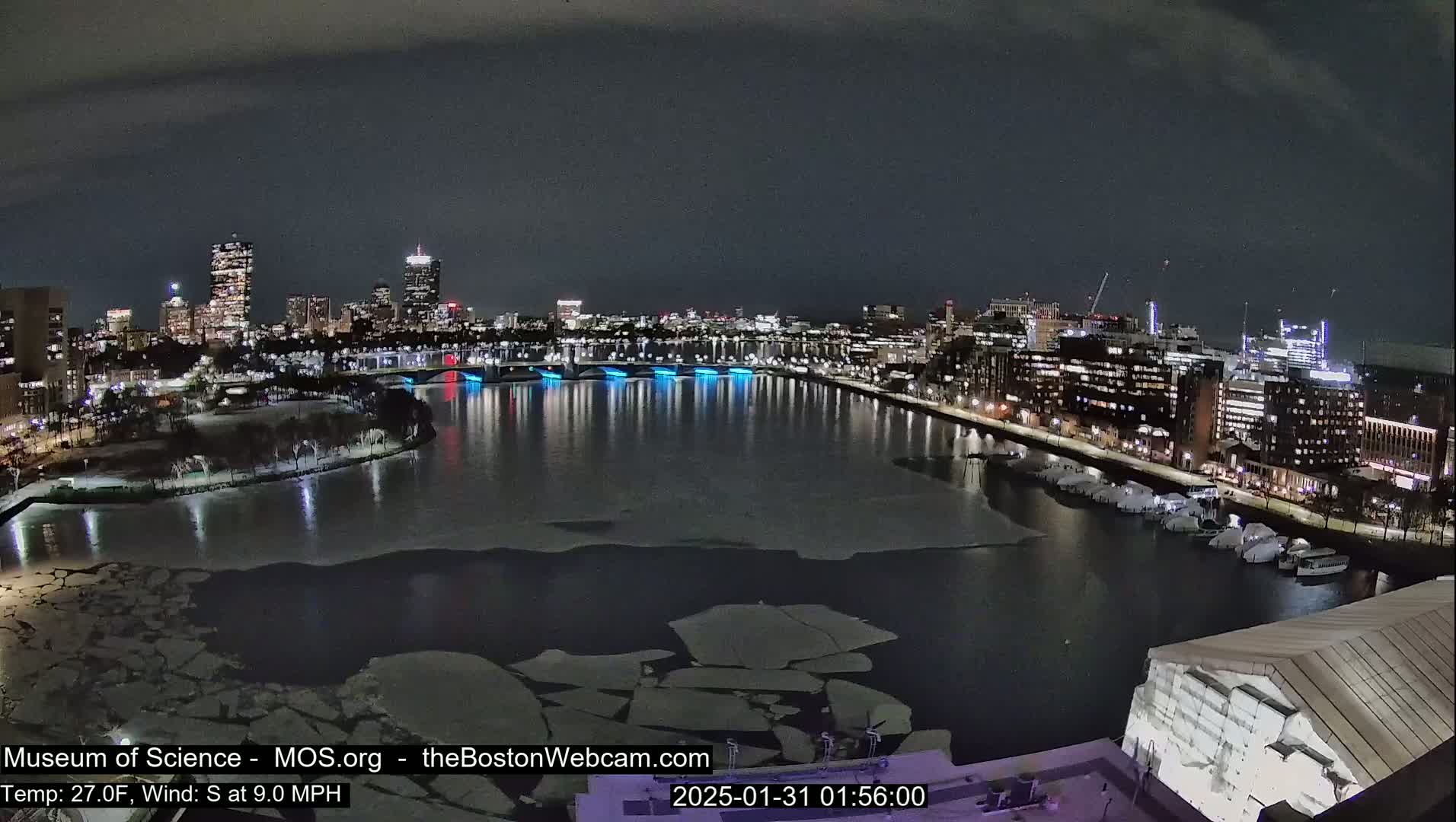 A nighttime view of a city skyline across a partially frozen river, with a south wind at 9 mph and a temperature of 27°F.