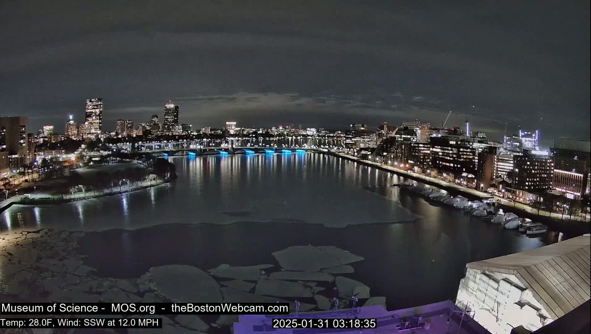 A nighttime cityscape with a river partially covered in ice, under a mostly cloudy sky.