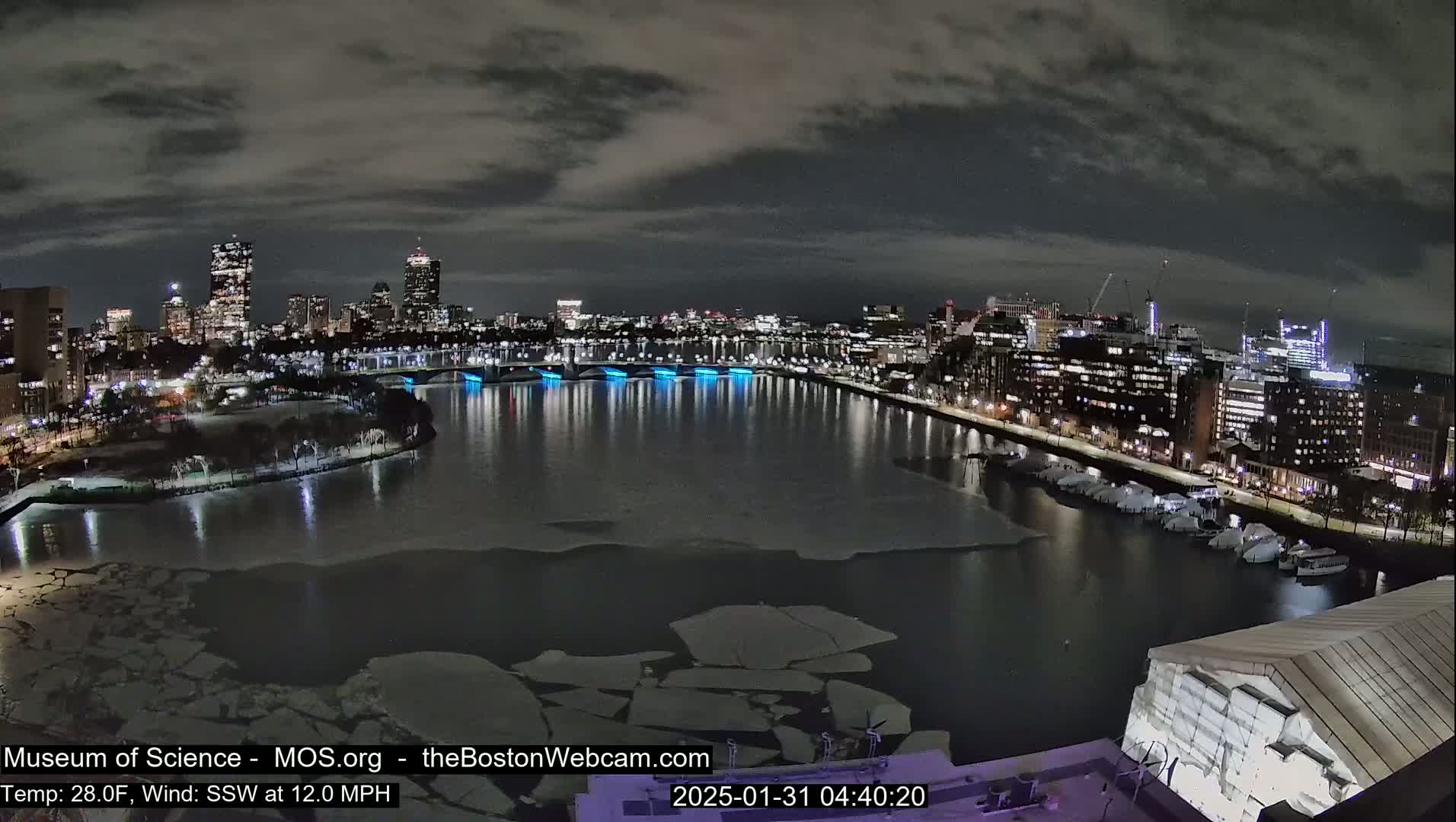 A nighttime view of a city skyline across a partially ice-covered river under a cloudy sky.