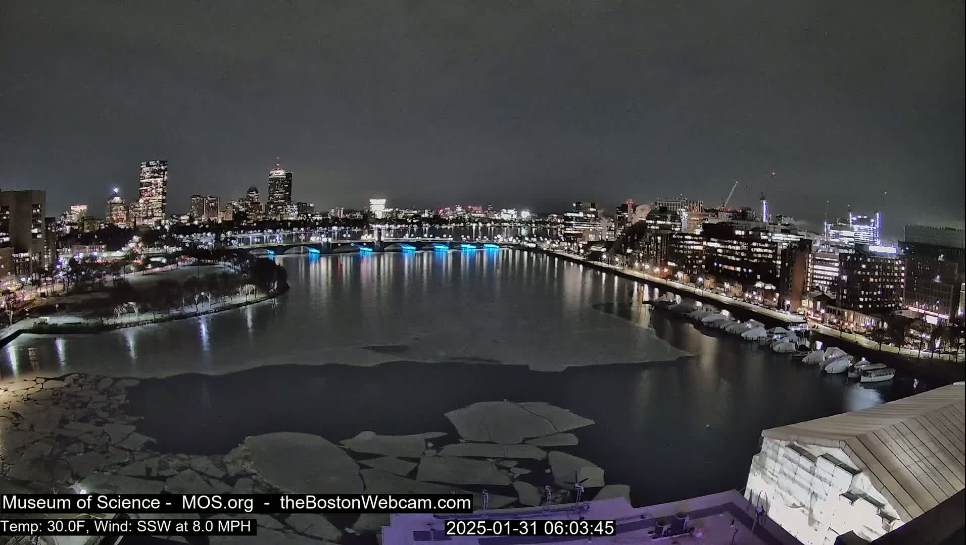 A nighttime view of a city skyline across a partially frozen river, under a dark sky.
