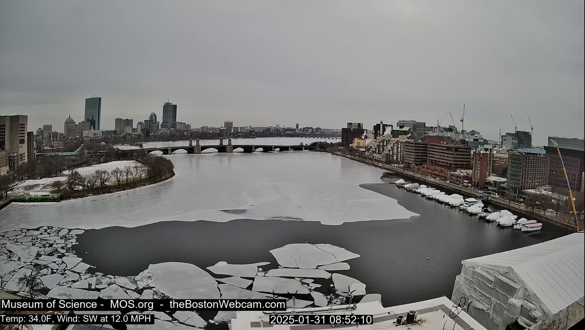 A partially frozen river flows through a city, with ice floes scattered across its surface under an overcast sky.