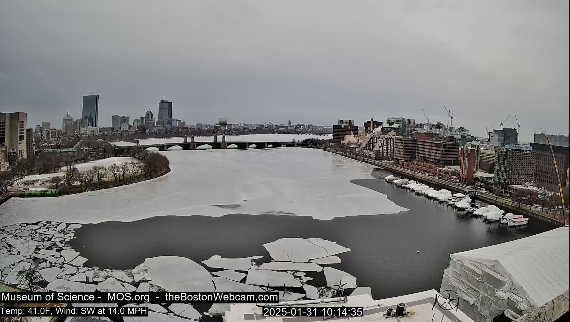 A partly frozen river flows past a city skyline under an overcast sky.