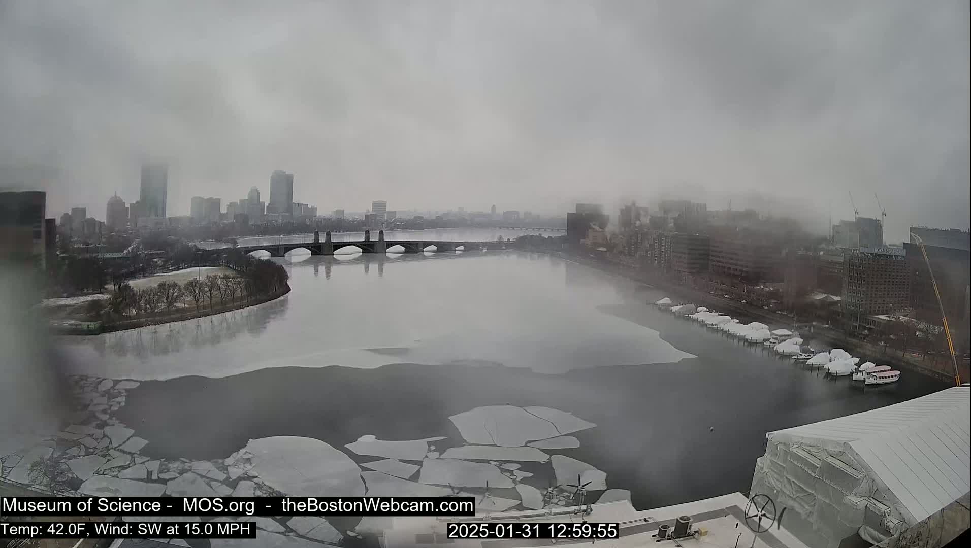 A hazy, cold day shows a city skyline across a river partially covered with ice floes, boats at a dock, and a bridge.