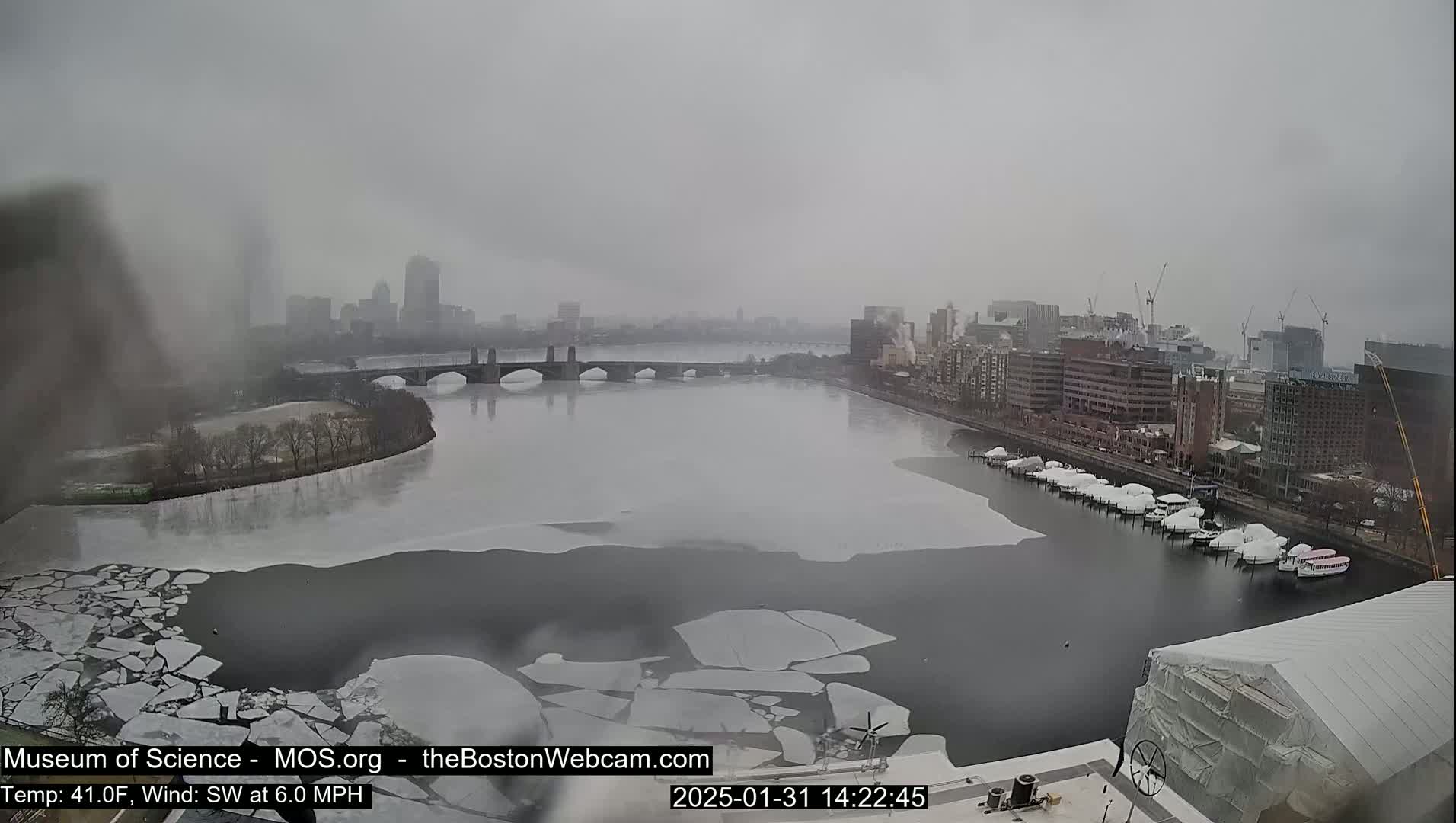 A partially frozen river flows past a city skyline under an overcast sky.