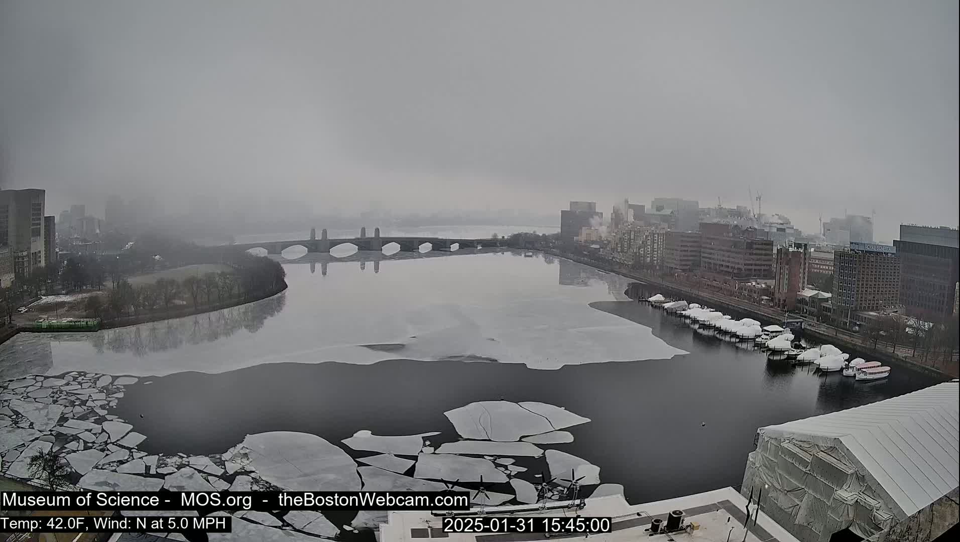 A foggy cityscape is seen from above, showing a river partially covered in ice with boats docked along the shore, and a bridge in the background.