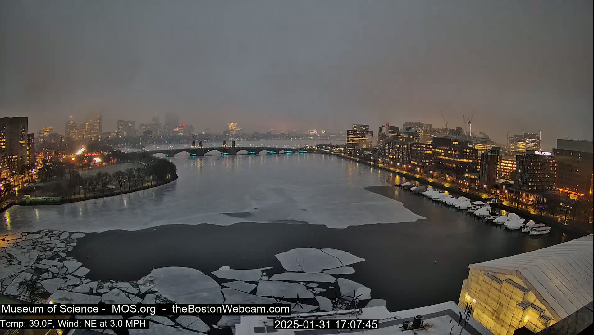 A nighttime cityscape with a river partially covered in ice floes under an overcast sky.