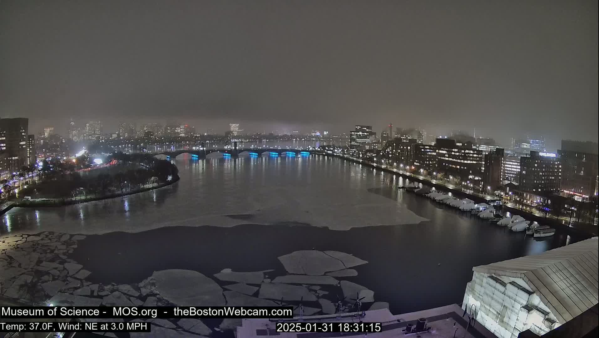 A nighttime view of a city waterfront with a partially ice-covered river, a bridge with blue lights, and docked boats under a hazy sky.