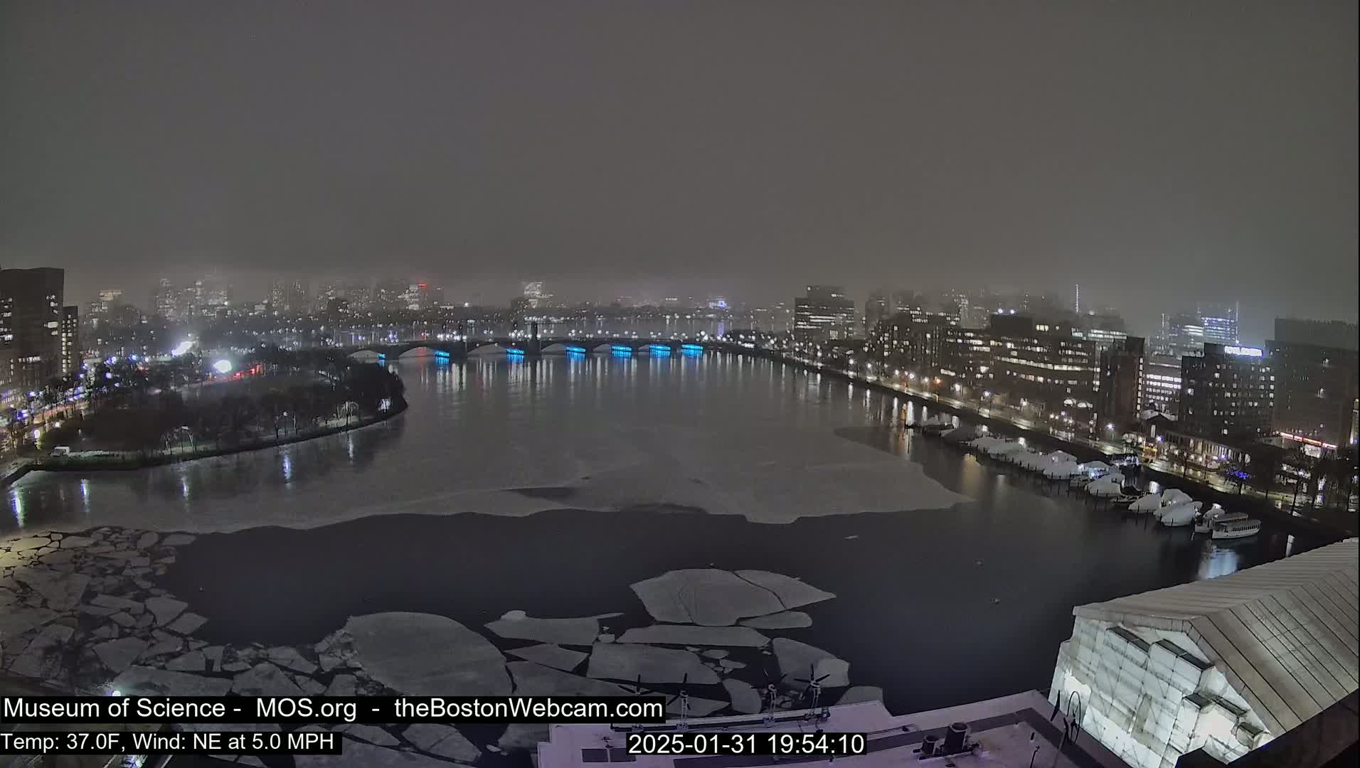A nighttime view of a city waterfront with a partially frozen river, under snowy conditions.