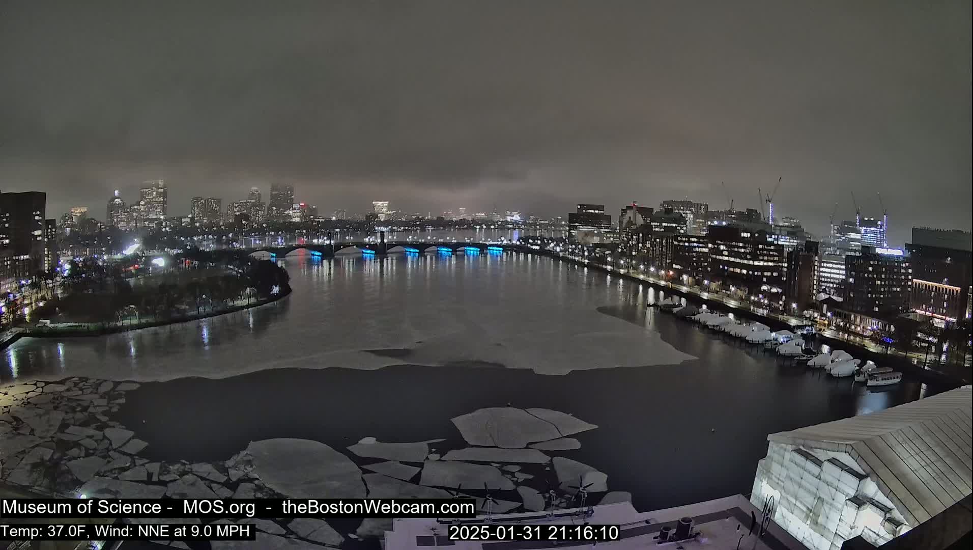 A nighttime cityscape with a river partially covered in ice, under a cloudy sky.