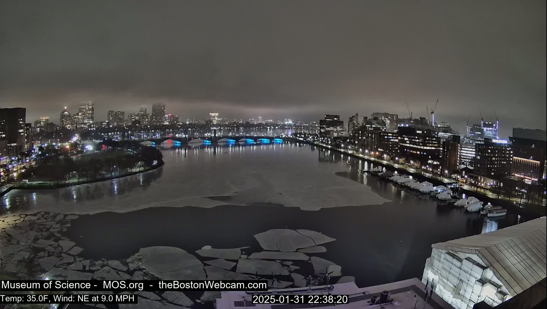 A nighttime cityscape with a partially frozen river, featuring a bridge illuminated in blue, under a foggy sky.