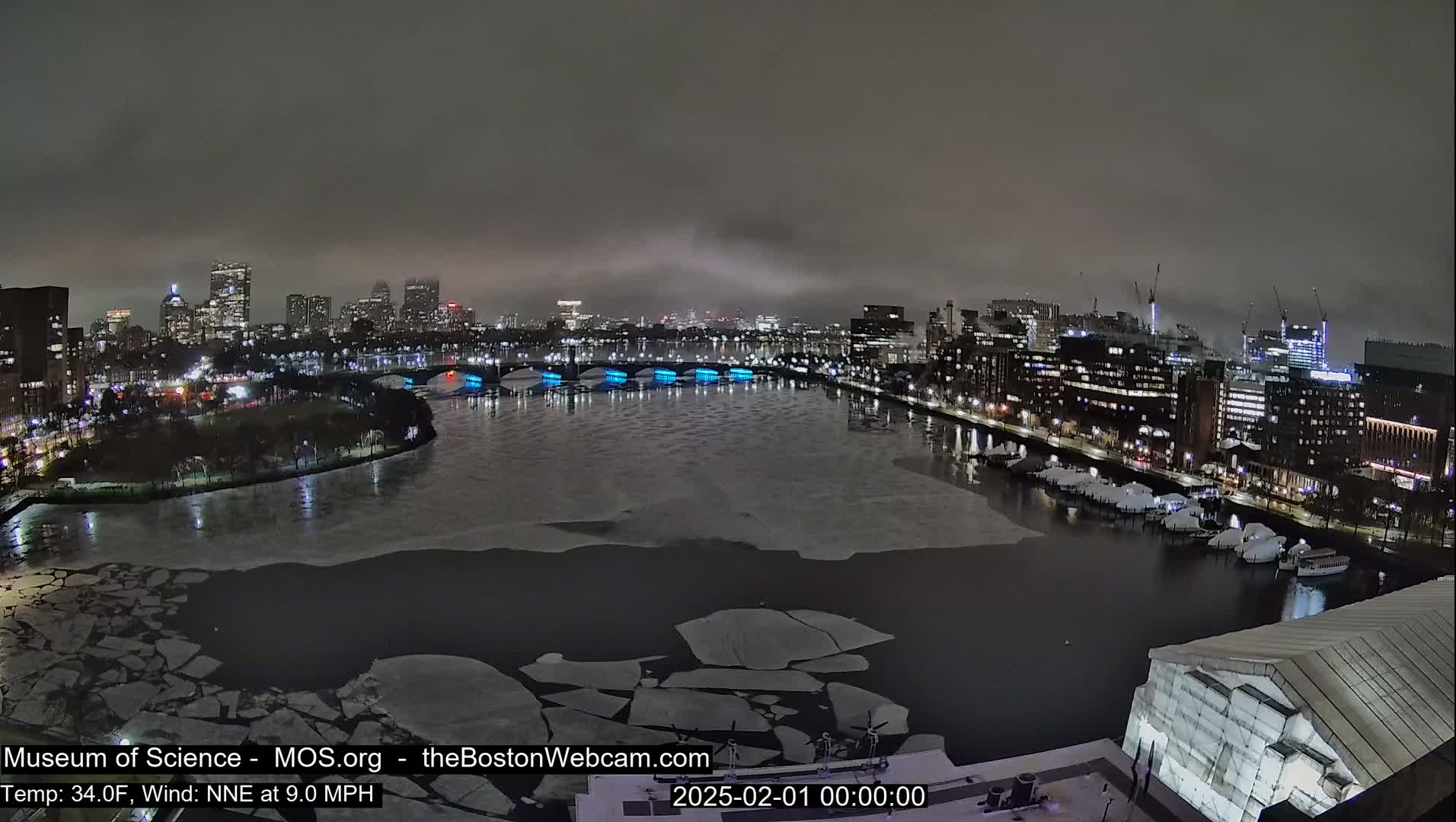 A nighttime view of a city skyline across a partially frozen river under a cloudy sky.