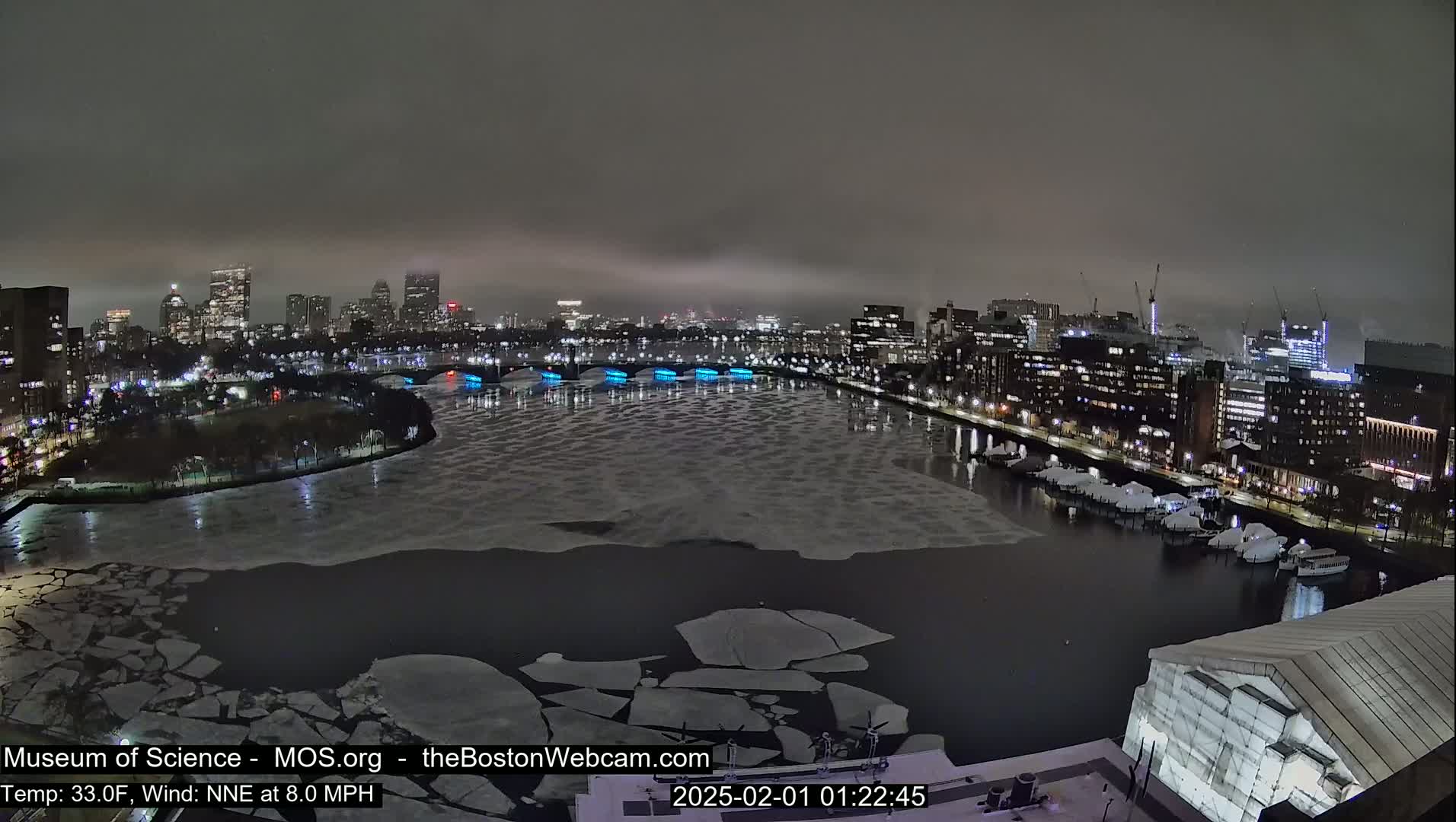 A nighttime cityscape with a partially frozen river, under a cloudy sky.