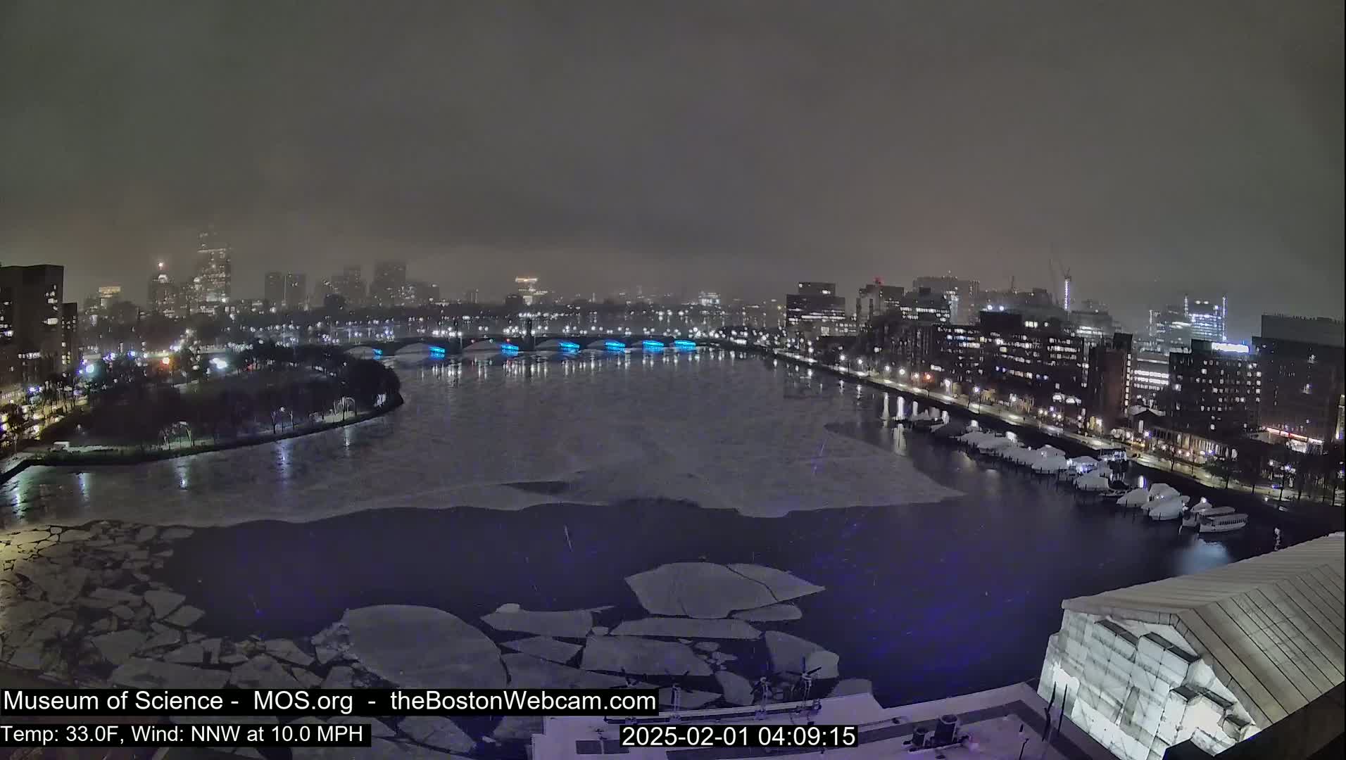 A nighttime cityscape with a partially frozen river, showing ice floes and city lights under a hazy sky.