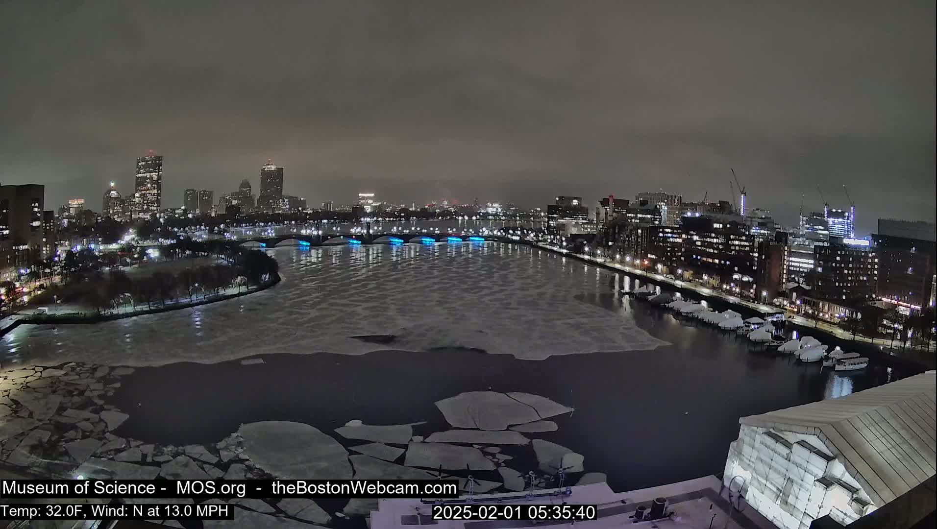 A nighttime view of a city skyline across a partially frozen river with a north wind at 13 miles per hour and a temperature of 32 degrees Fahrenheit.