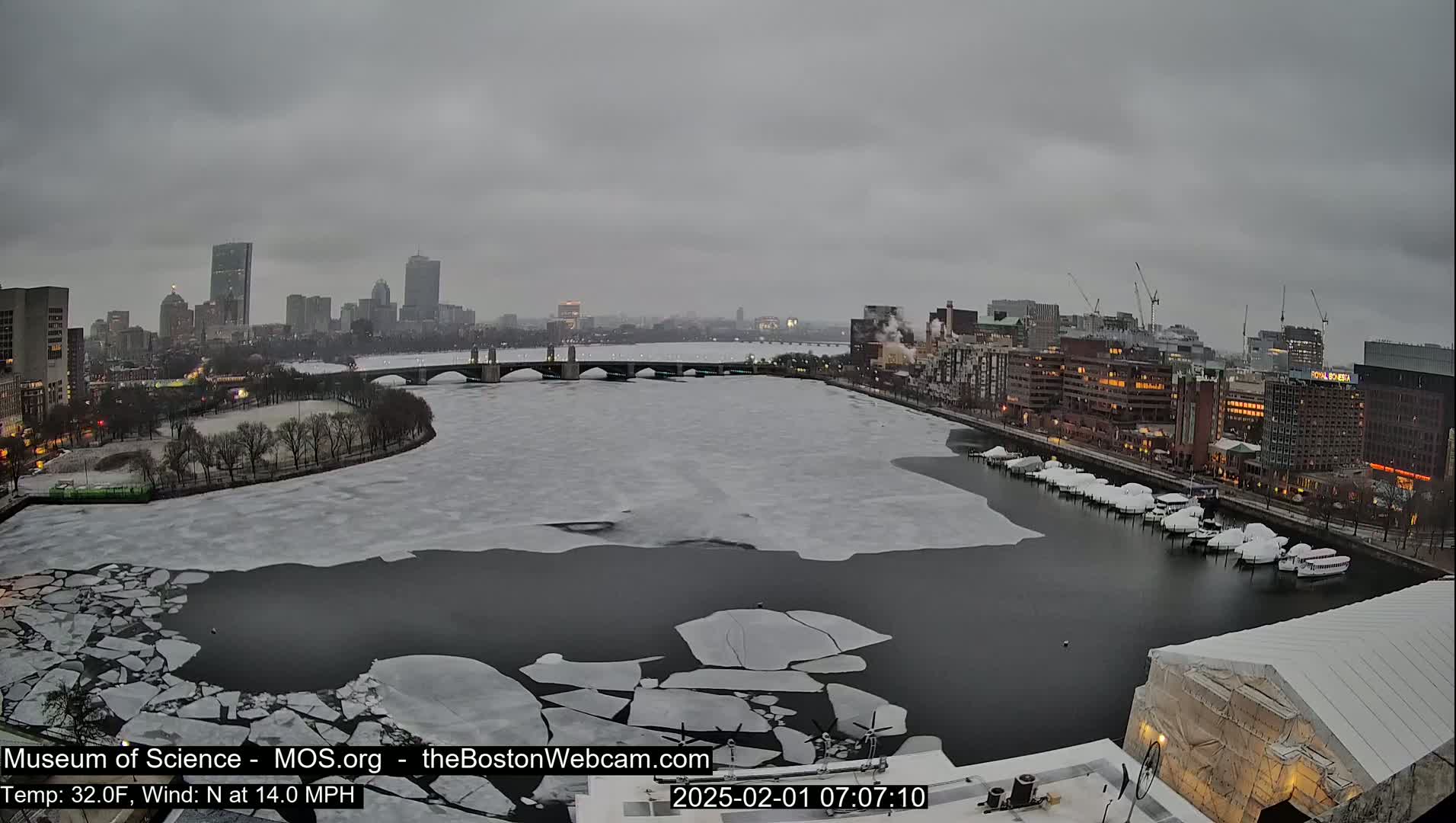 A city skyline is visible across a partially frozen river under an overcast sky.