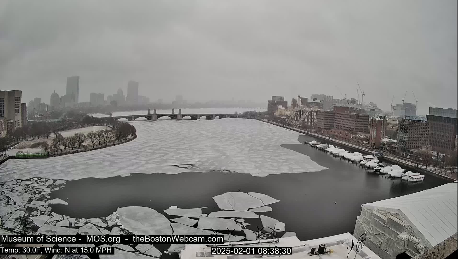A partly frozen river flows past a city skyline under a snowy, overcast sky.