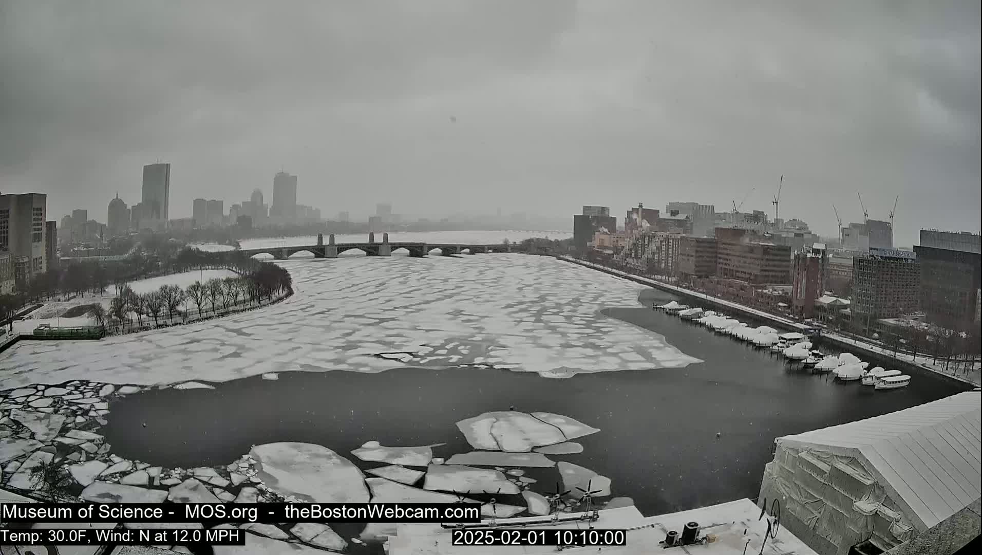 A snow-covered city skyline is seen across a partially frozen river with boats docked along the shore in snowy conditions.