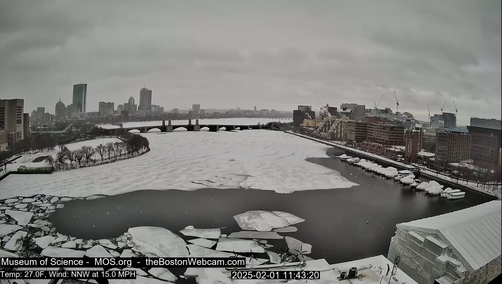 A snow-covered city skyline is visible across a partially frozen river, with a bridge in the mid-ground and a snow-covered structure in the foreground, under overcast skies.