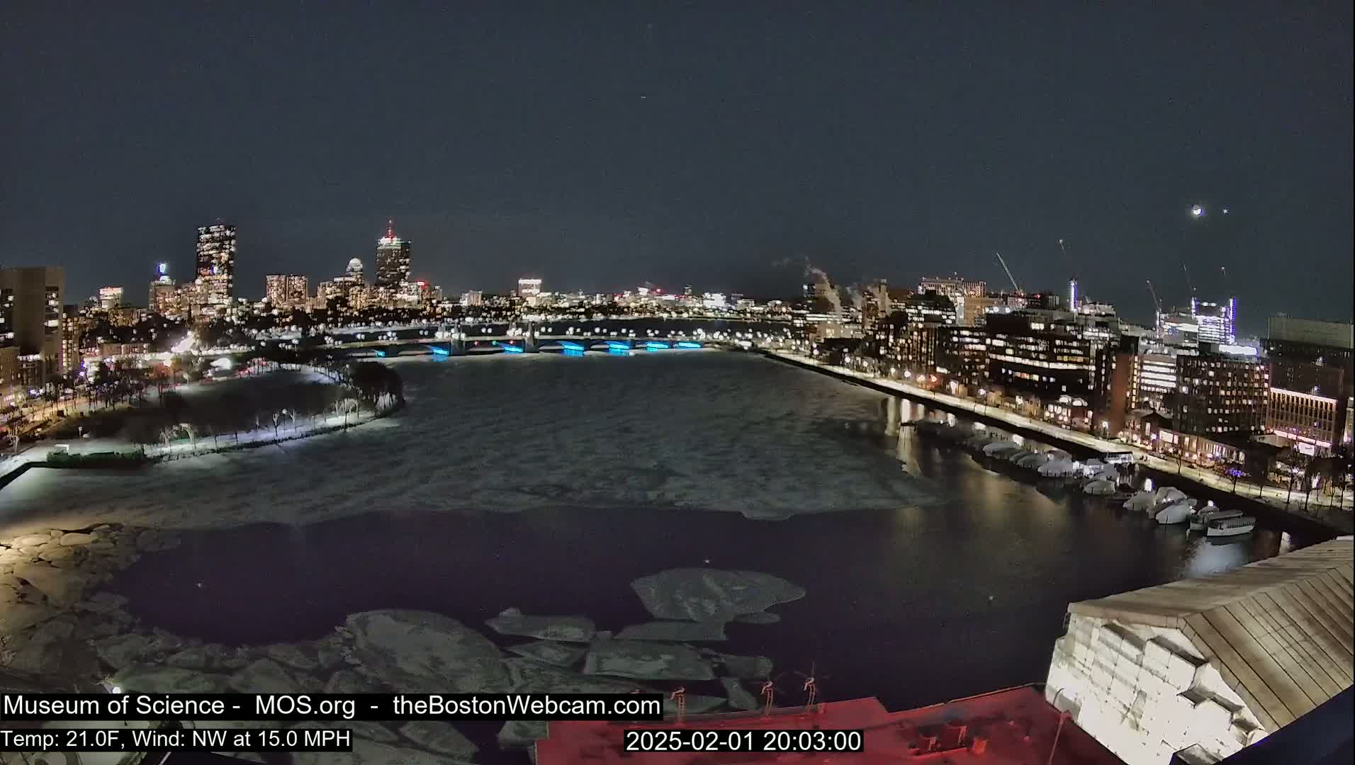 A nighttime cityscape with a partially frozen river, illuminated buildings, and a northwest wind at 15 mph in 21-degree Fahrenheit weather.