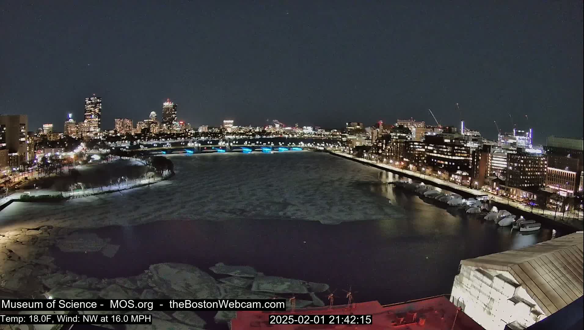 A nighttime view of a city skyline across a partially ice-covered river, with a northwest wind at 16 mph and a temperature of 18°F.