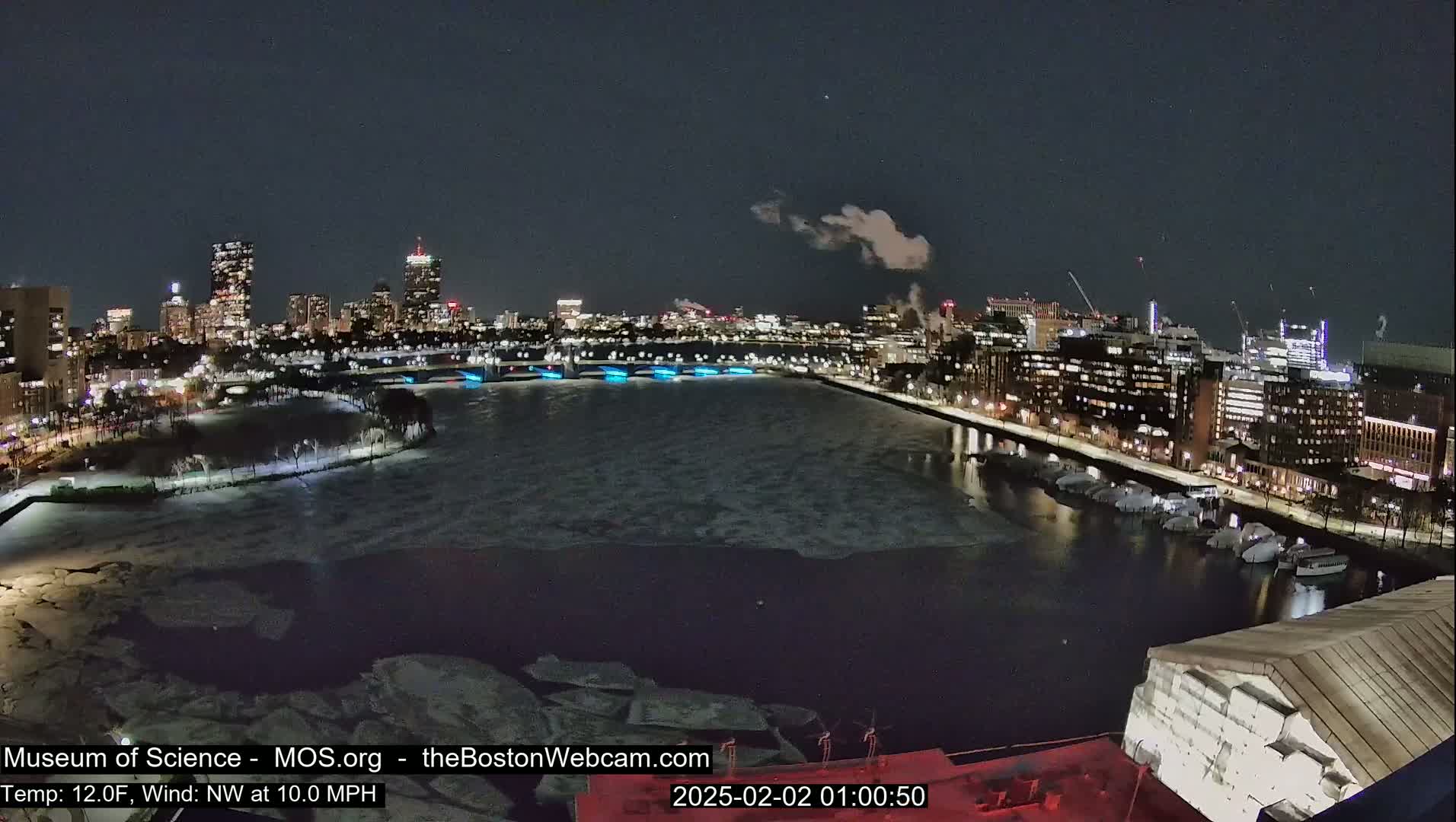 A nighttime view of a city skyline across a partially ice-covered river, with a 10 mph northwest wind and a temperature of 12 degrees Fahrenheit.